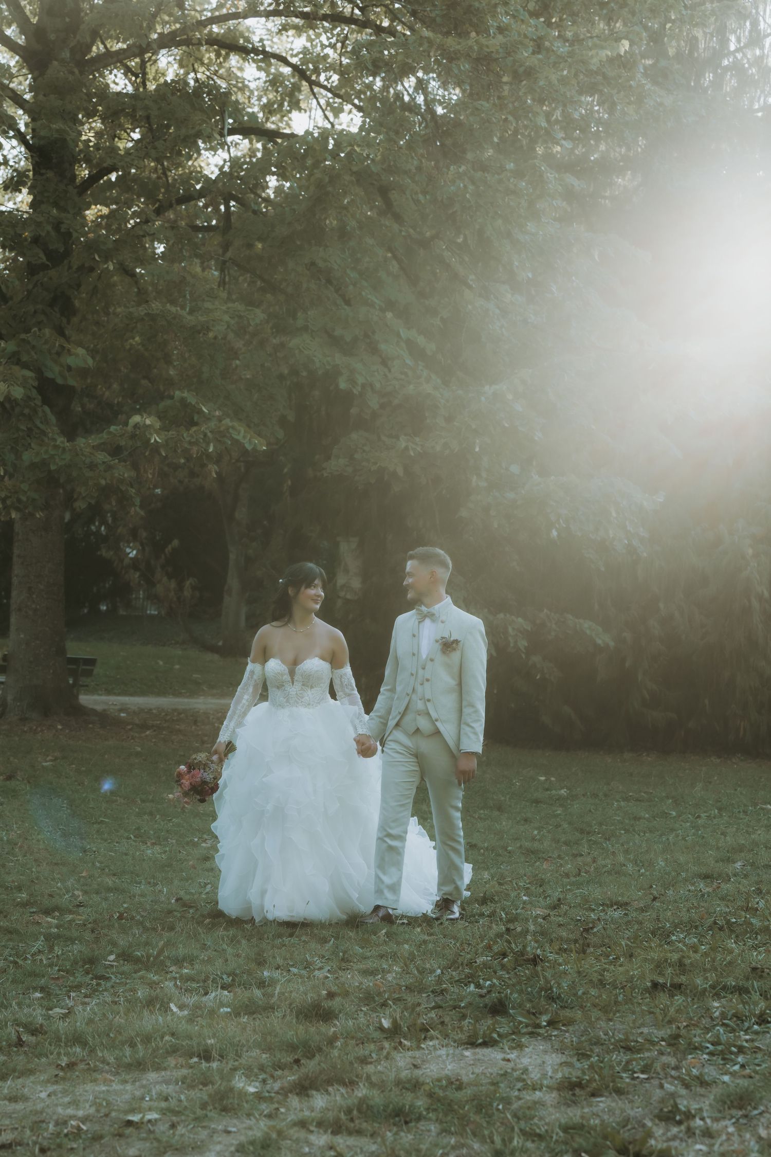 Bride and groom walk hand-in-hand through a sunlit park, she in a white gown, he in a light suit.