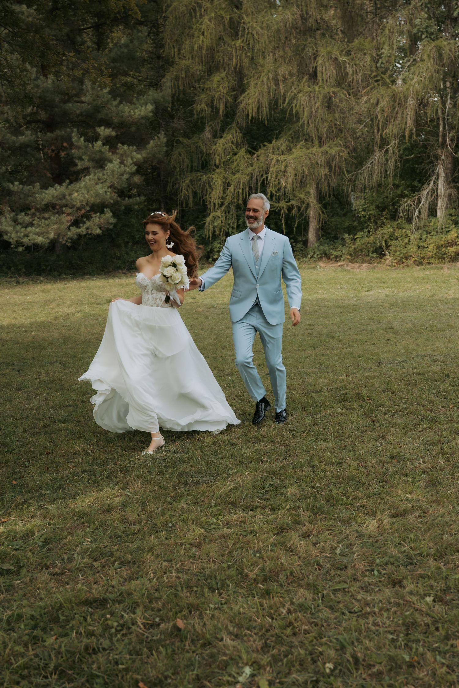 Bride in white gown and groom in light blue suit run hand-in-hand across green lawn with trees behind them.