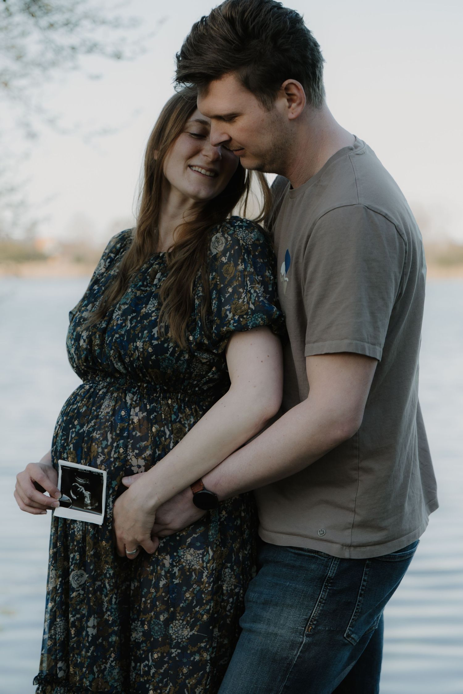 Pregnant couple holding ultrasound photo by a lake, woman in floral dress smiling as man tenderly kisses her head.