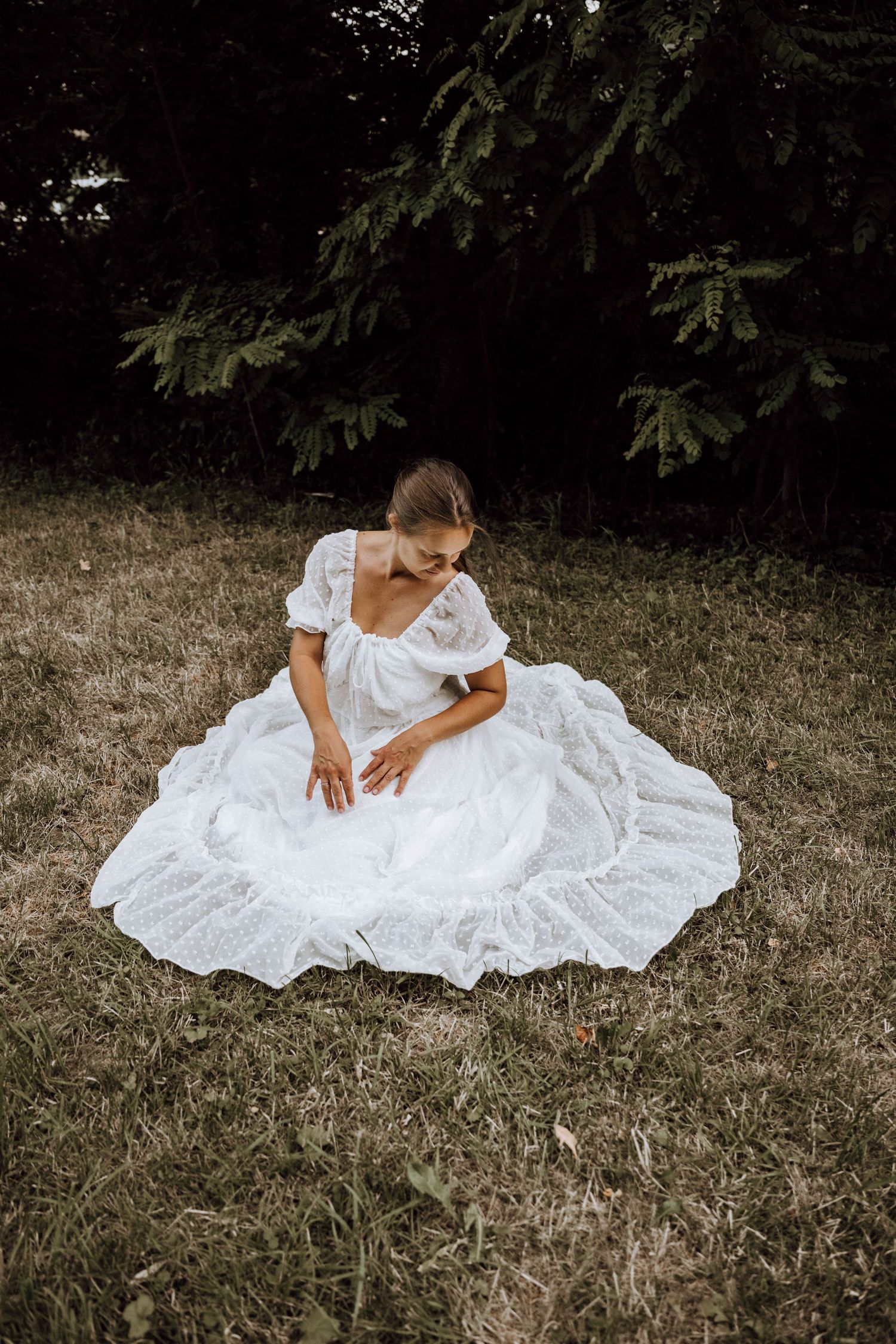 A bride in a flowing white ball gown sits gracefully on grass surrounded by lush green trees.