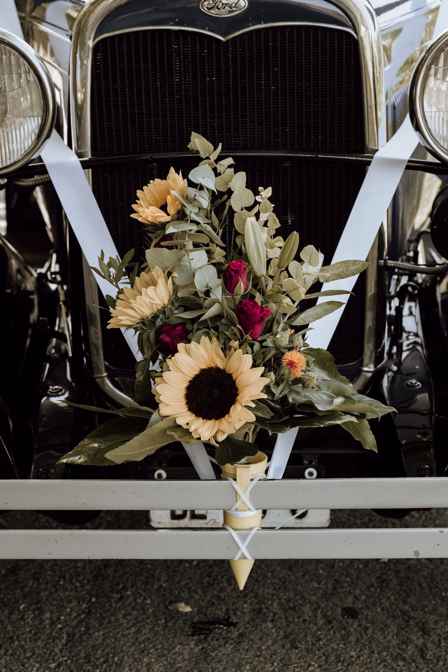 Vintage Ford wedding car decorated with sunflowers, red roses, and eucalyptus bouquet tied with white ribbon on the front grille.