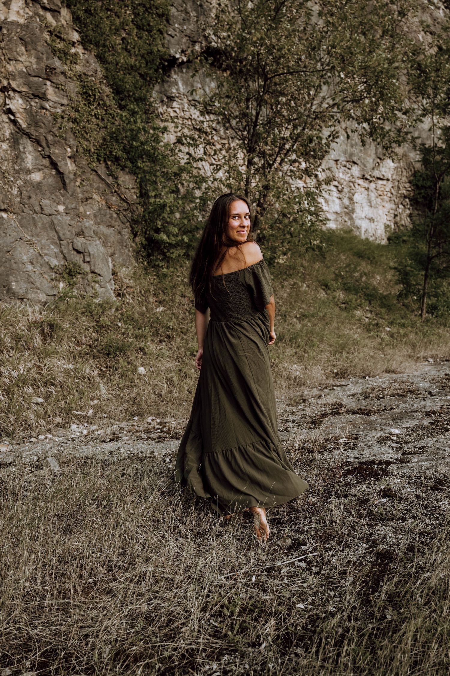 Woman in olive green off-shoulder maxi dress standing in a rocky, wooded outdoor setting with long dark hair.