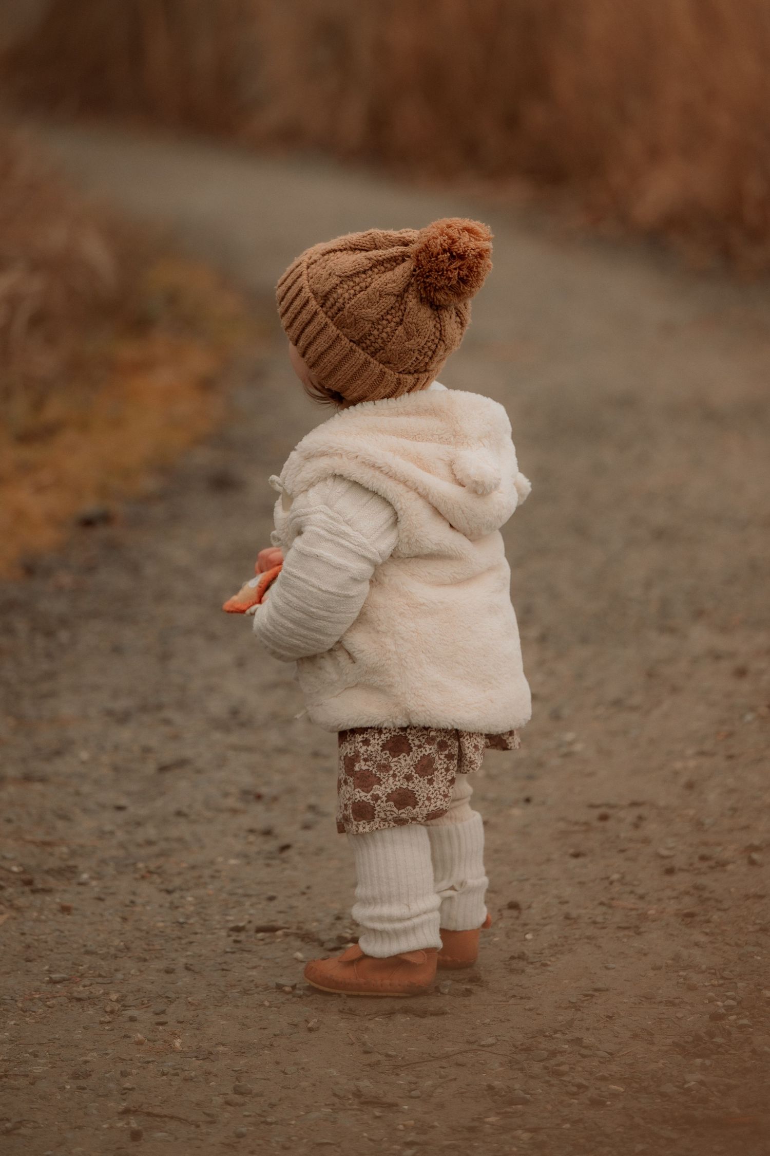Toddler in white fur coat, knit pom-pom hat, and leg warmers standing on a dirt path in autumn.