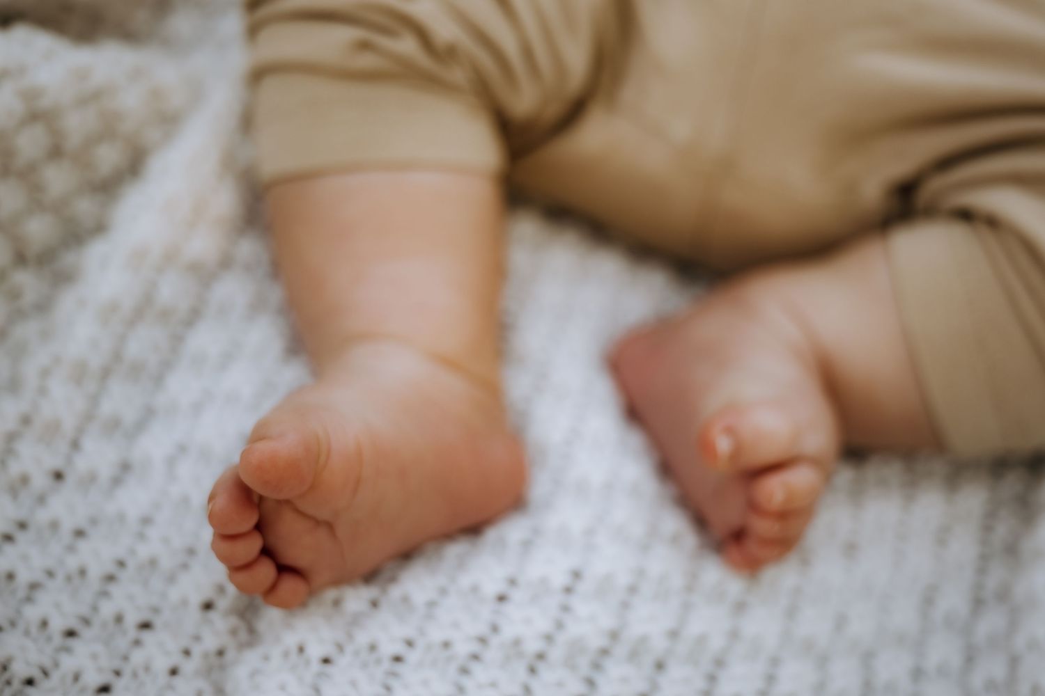 Close-up of tiny newborn baby feet resting on a soft white knit blanket, wearing a beige onesie.