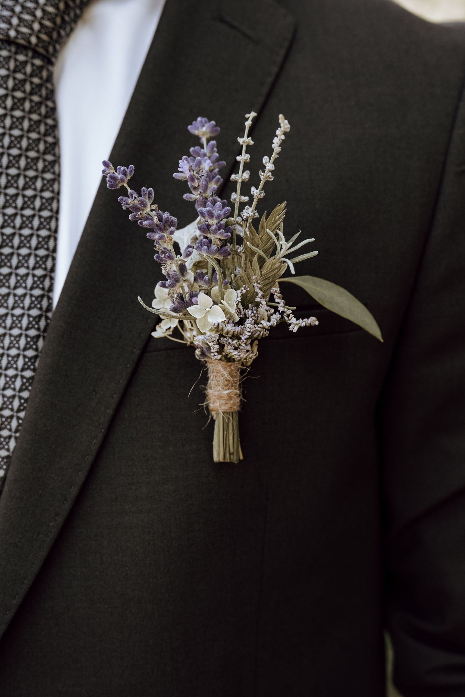 A rustic boutonniere featuring purple lavender and white flowers tied with twine, pinned to a dark suit lapel.