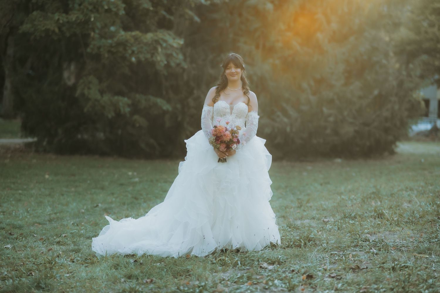 Bride in a flowing white lace wedding gown holding a colorful bouquet, standing in a sunlit garden.