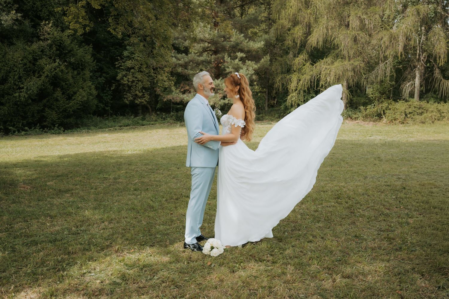 Bride in flowing white gown and groom in light blue suit share a romantic dance outdoors on lush green grass.