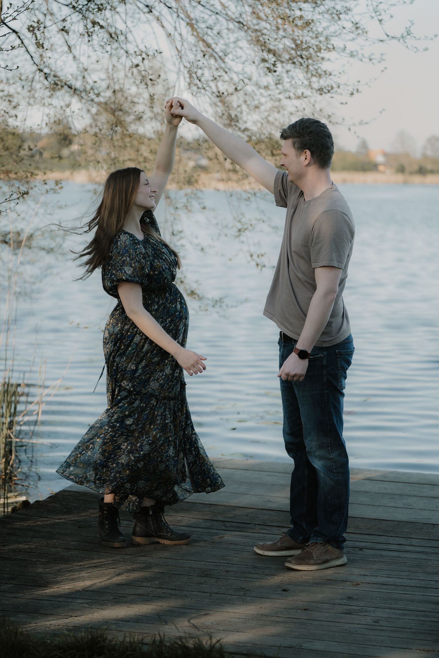 Pregnant couple dancing on lakeside dock, man spinning woman in floral dress near blooming trees.