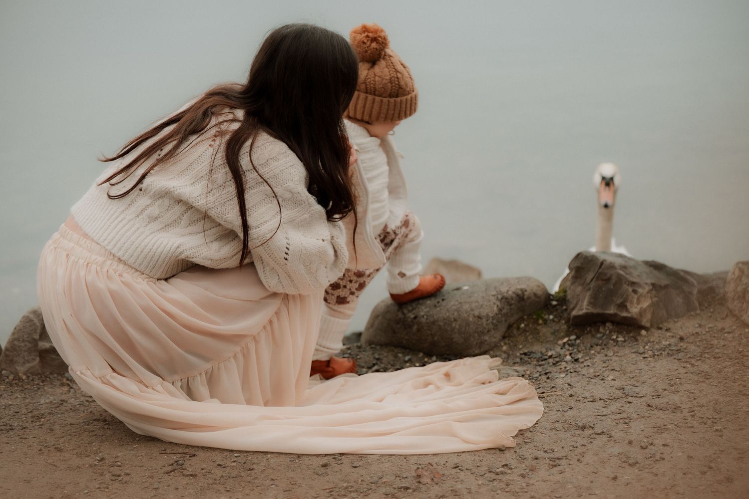 Mother and child crouching by a misty lakeside, watching a white swan, wearing cozy knit sweaters and a pom-pom hat.