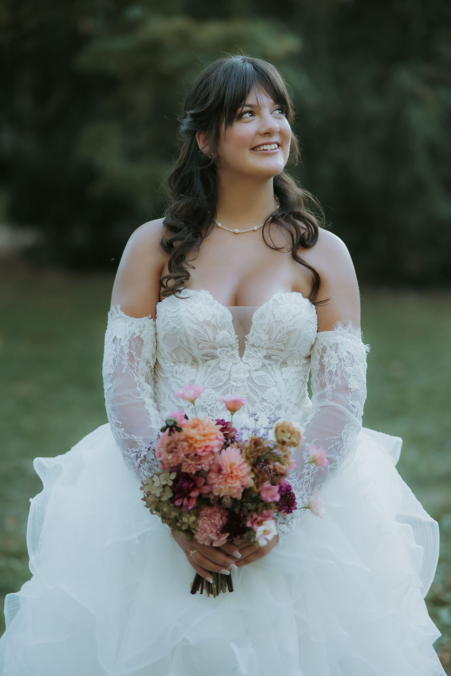 Smiling bride in off-shoulder lace wedding gown holding colorful bouquet of pink and purple flowers outdoors.