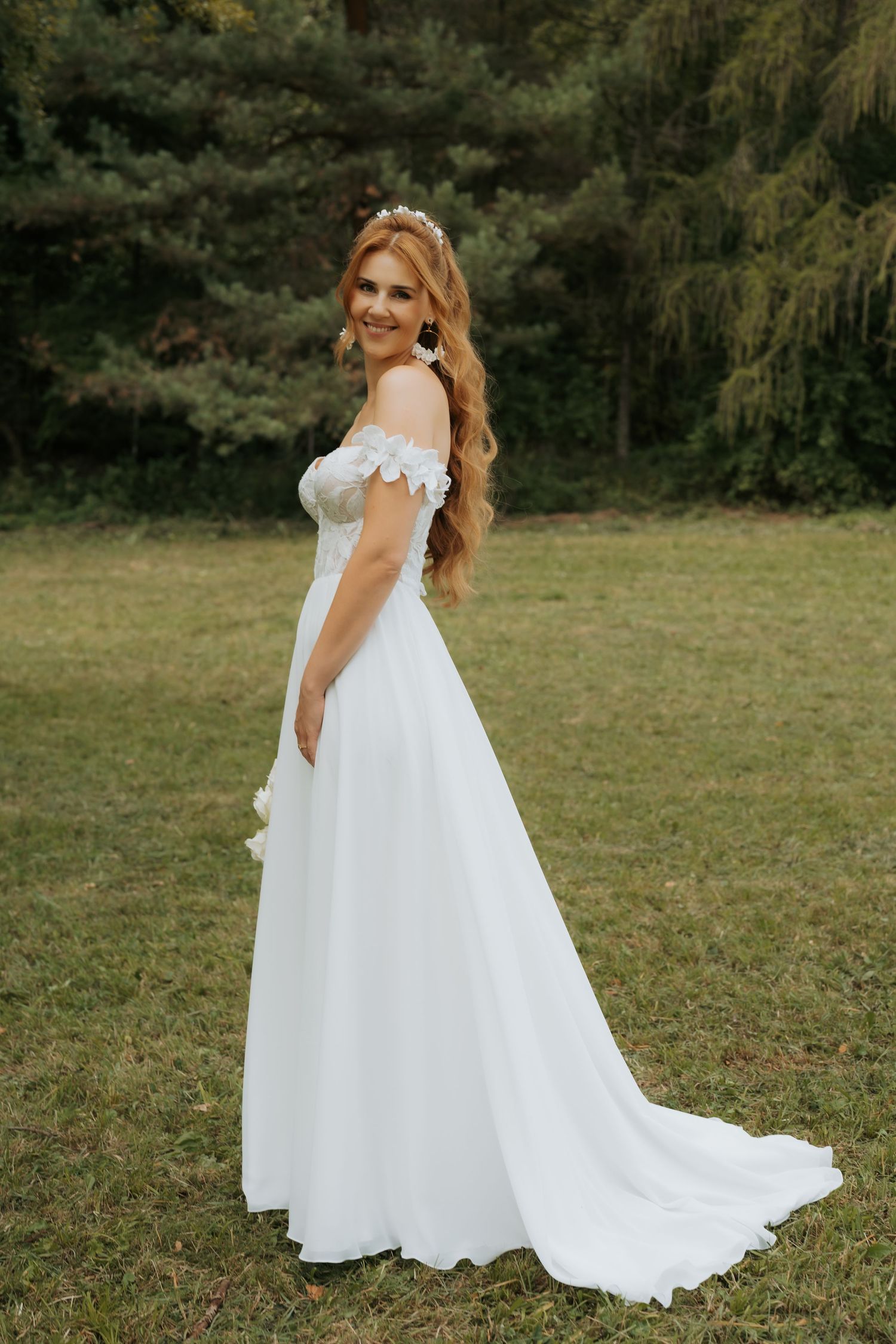 Smiling bride in off-shoulder white lace wedding gown with flowing train poses outdoors on green grass.