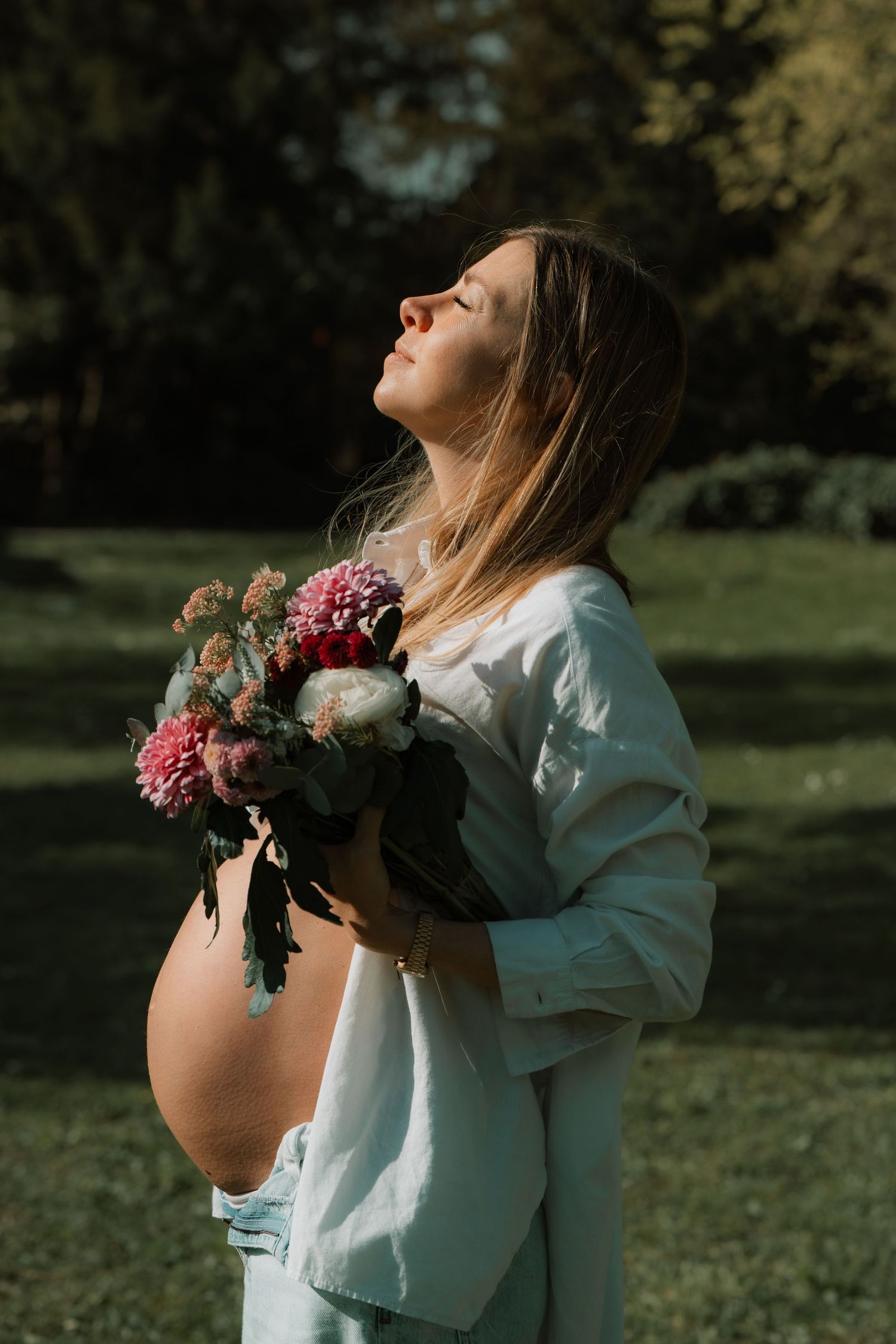 Pregnant woman in white shirt holds pink flower bouquet outdoors, head tilted back in sunlight with green trees behind.