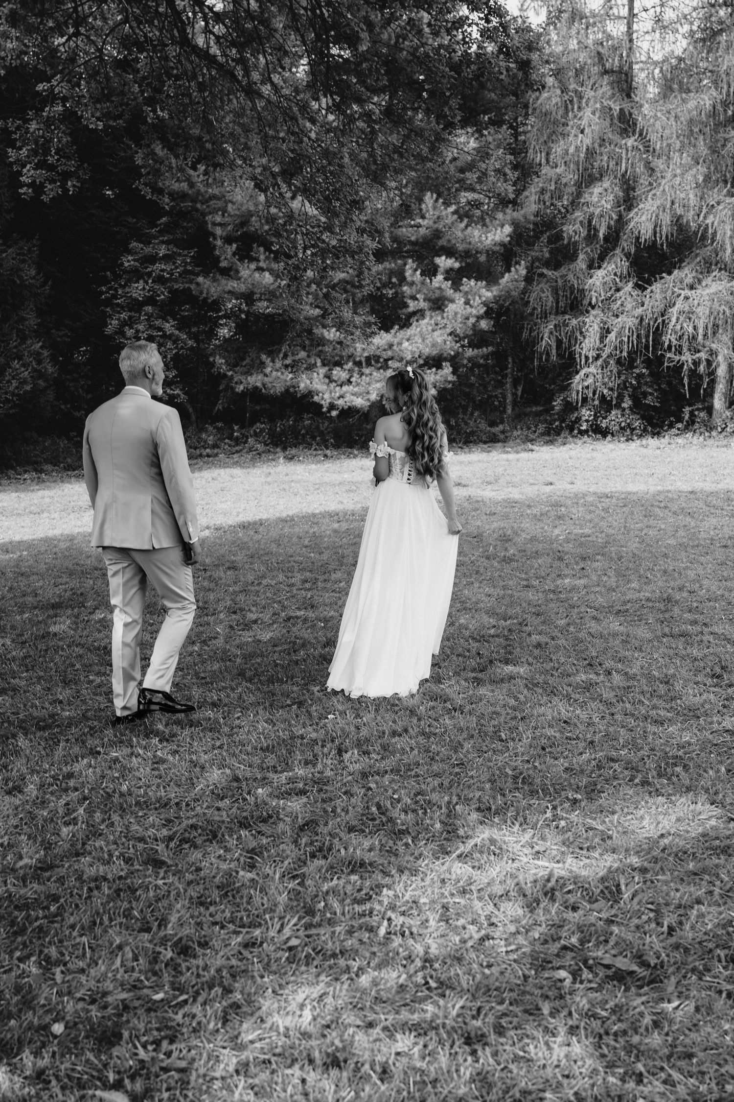Black and white photo of a bride and groom walking hand in hand across a grassy lawn toward lush trees.
