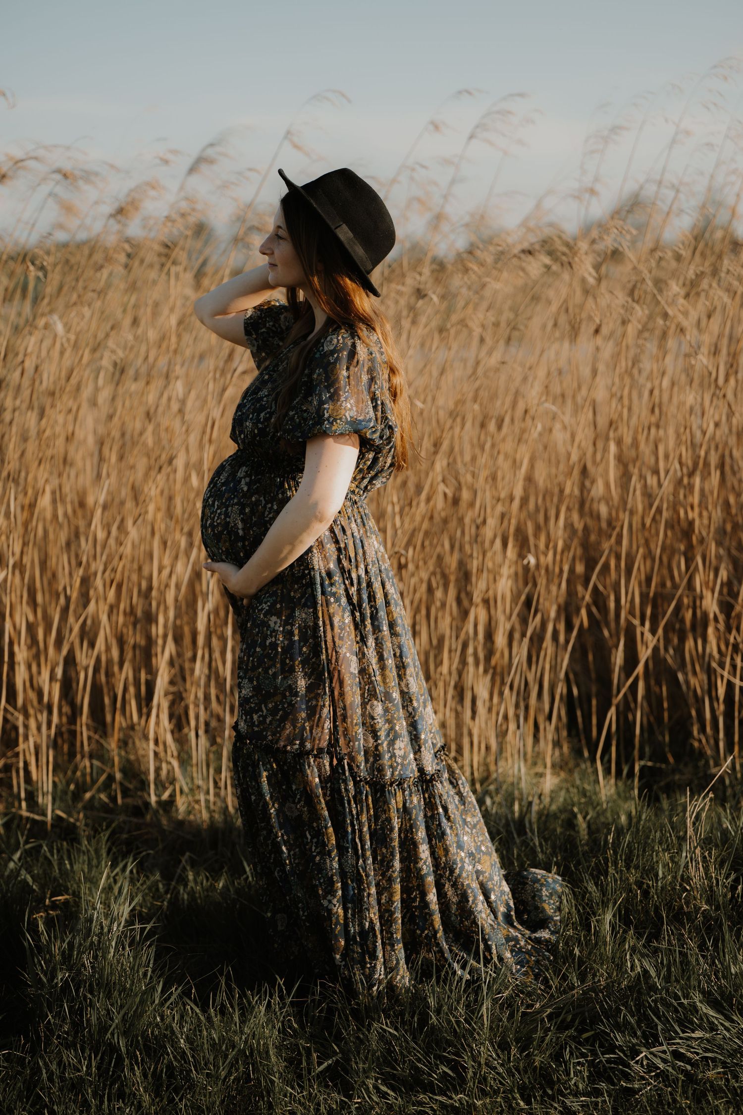 Pregnant woman in floral maxi dress and black hat standing in golden wheat field during golden hour.