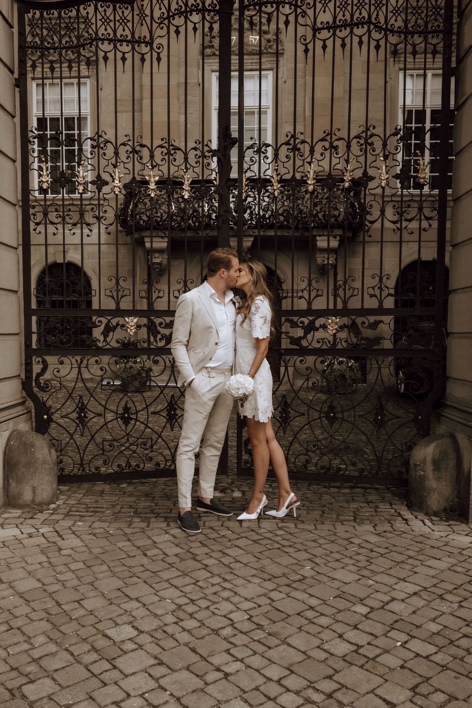 Couple sharing a romantic kiss in white wedding attire before ornate iron gates on cobblestone street.