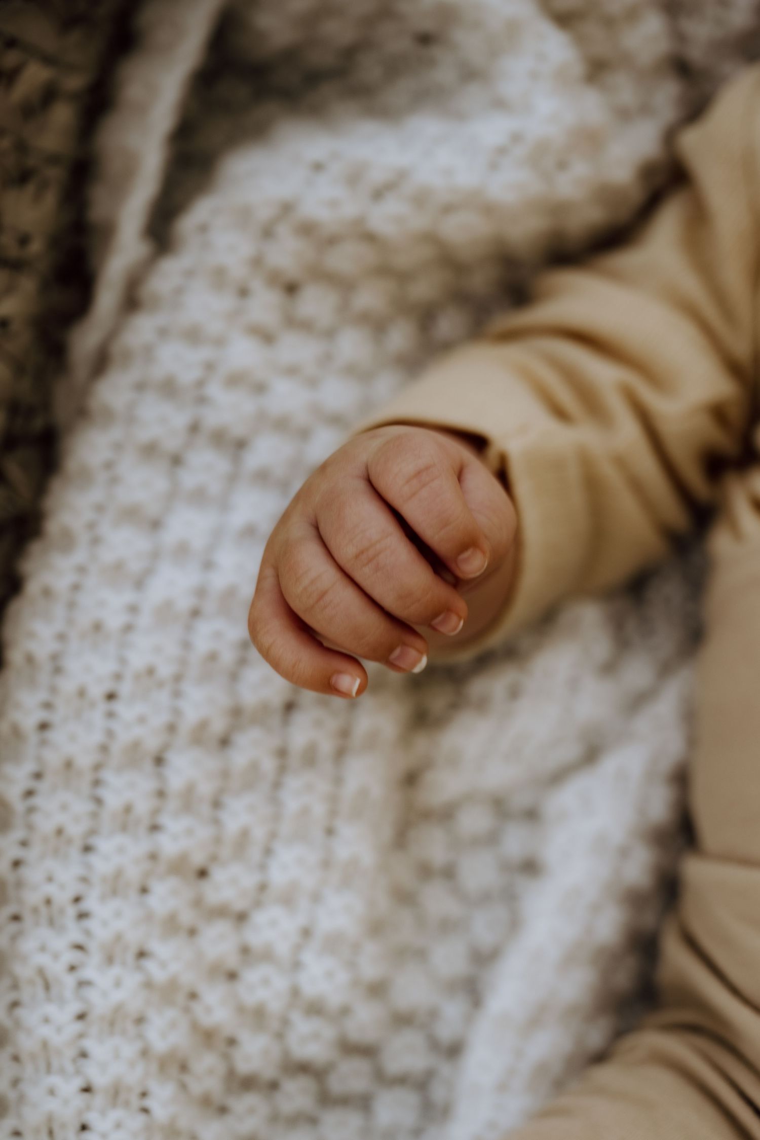 Close-up of a newborn baby's tiny hand resting on a soft white knit blanket, wearing a beige onesie.