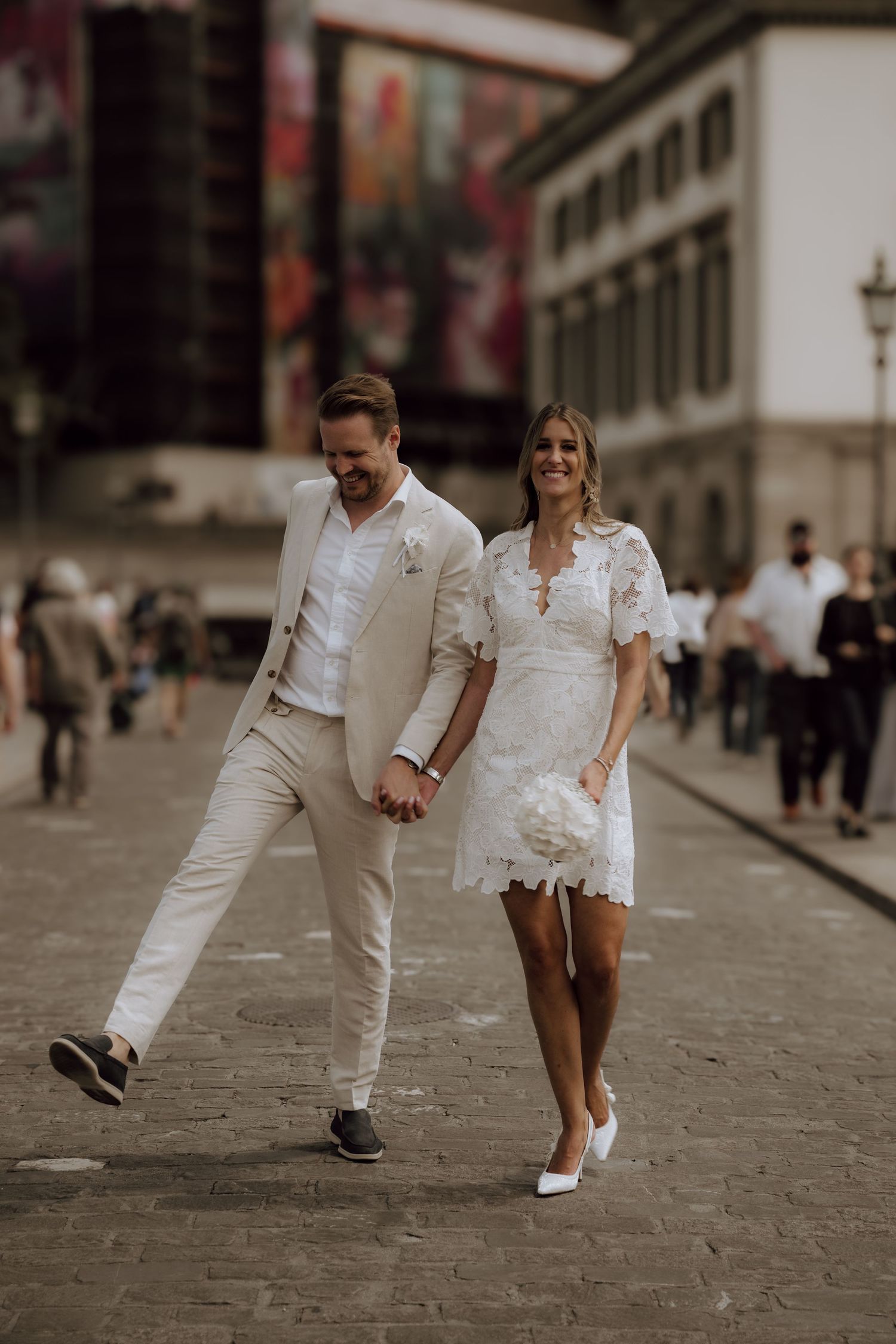 Couple in wedding attire walking hand-in-hand on cobblestone street, bride in white lace mini dress holding bouquet.