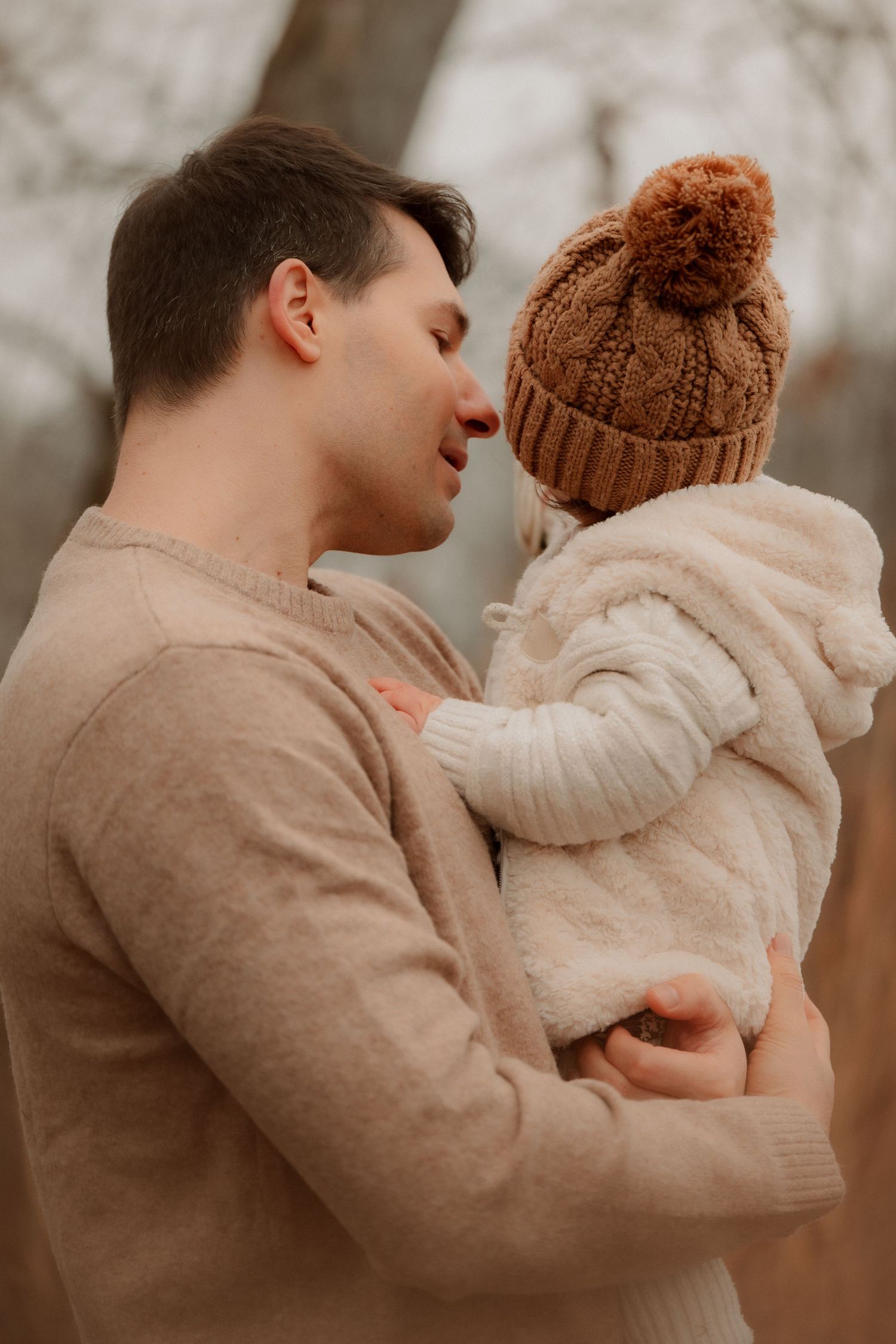A father tenderly holds his baby dressed in a white knit outfit and brown pom-pom hat outdoors in autumn.