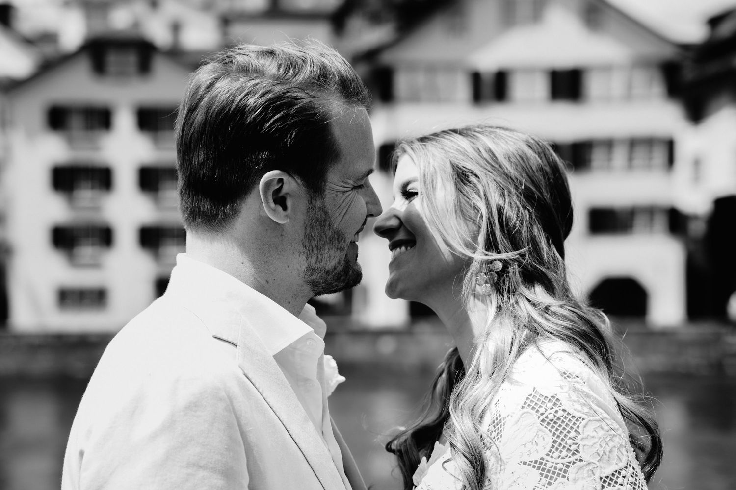Black and white photo of a romantic couple smiling and leaning close together outdoors near European buildings.