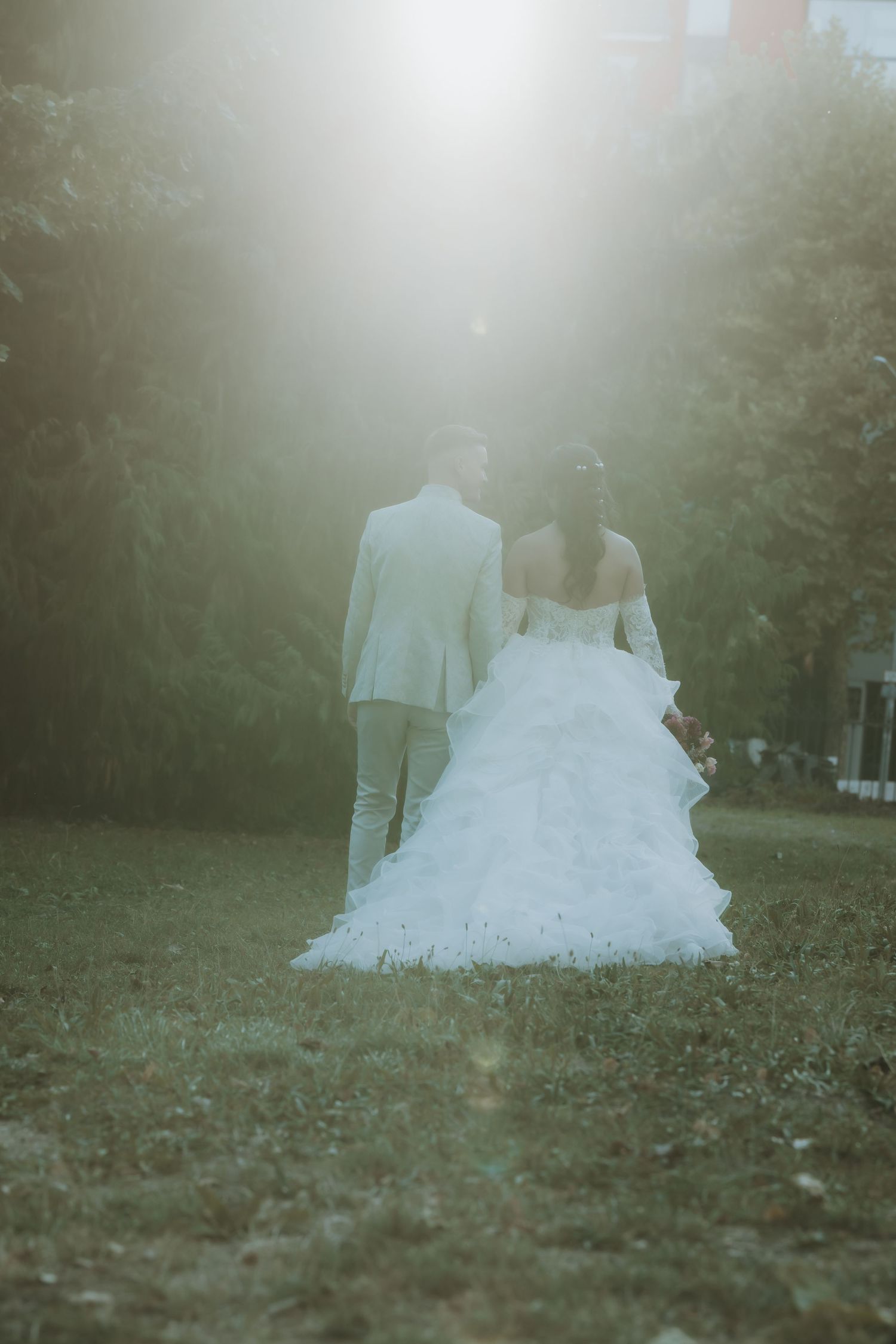 Bride and groom walking away in sunlit garden, bride wearing elegant white ruffled gown, groom in light suit.