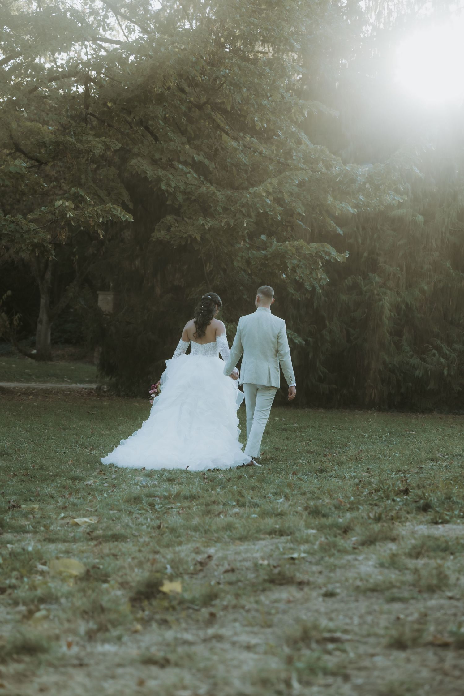 Bride in white gown and groom in light suit walking hand in hand through a sunlit green park.