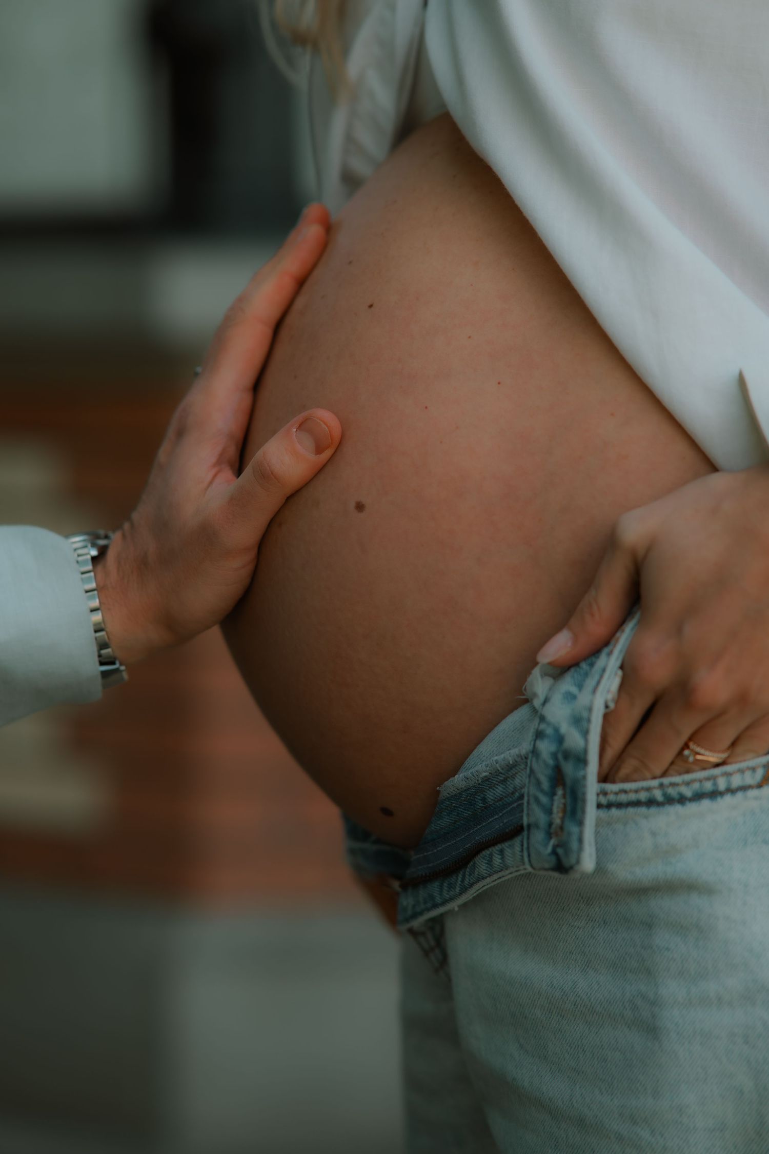 Close-up of a pregnant woman cradling her bare baby bump, wearing unbuttoned light blue jeans and a white shirt.