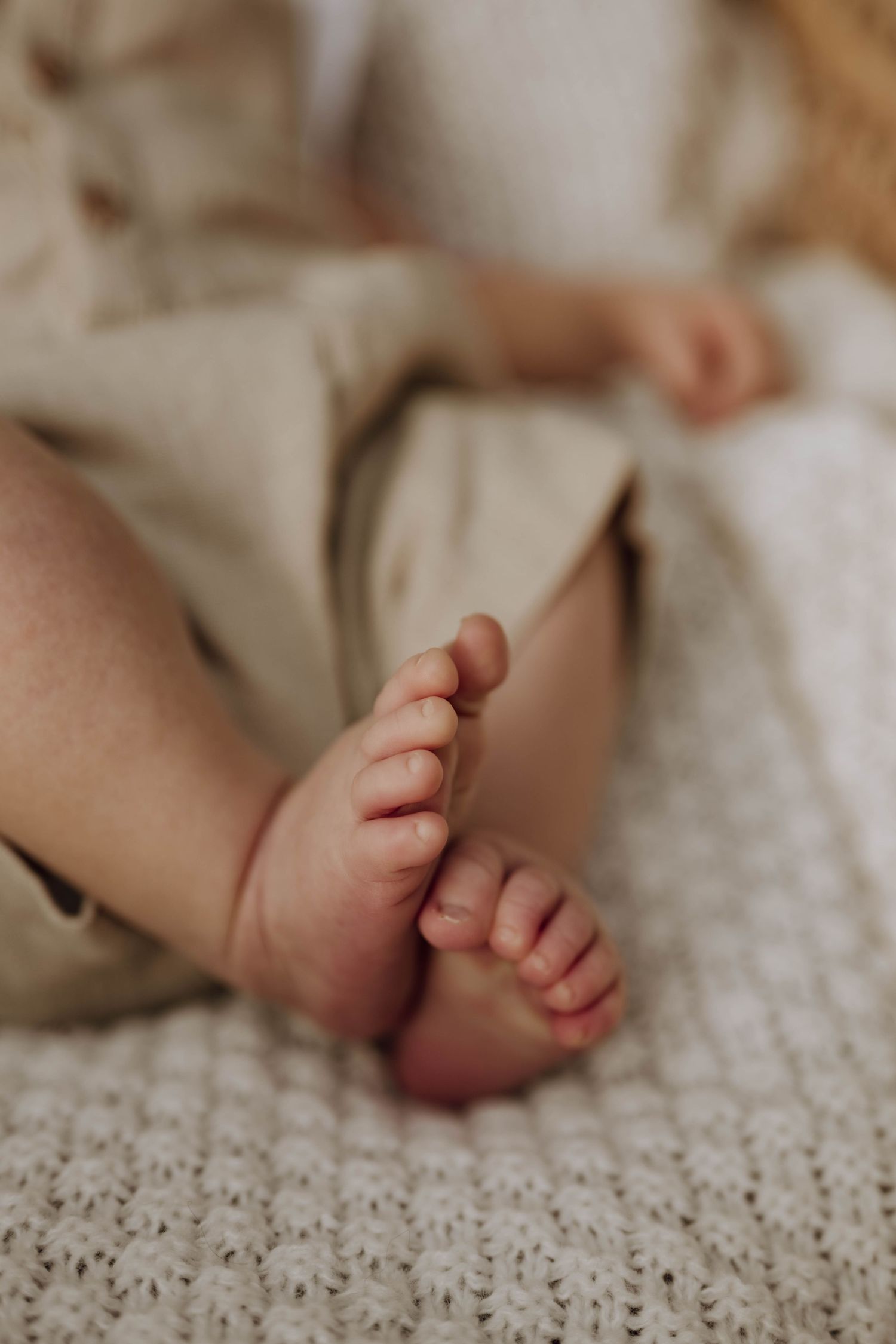 Close-up of tiny newborn baby feet resting on a soft white knit blanket, wearing beige linen pants.