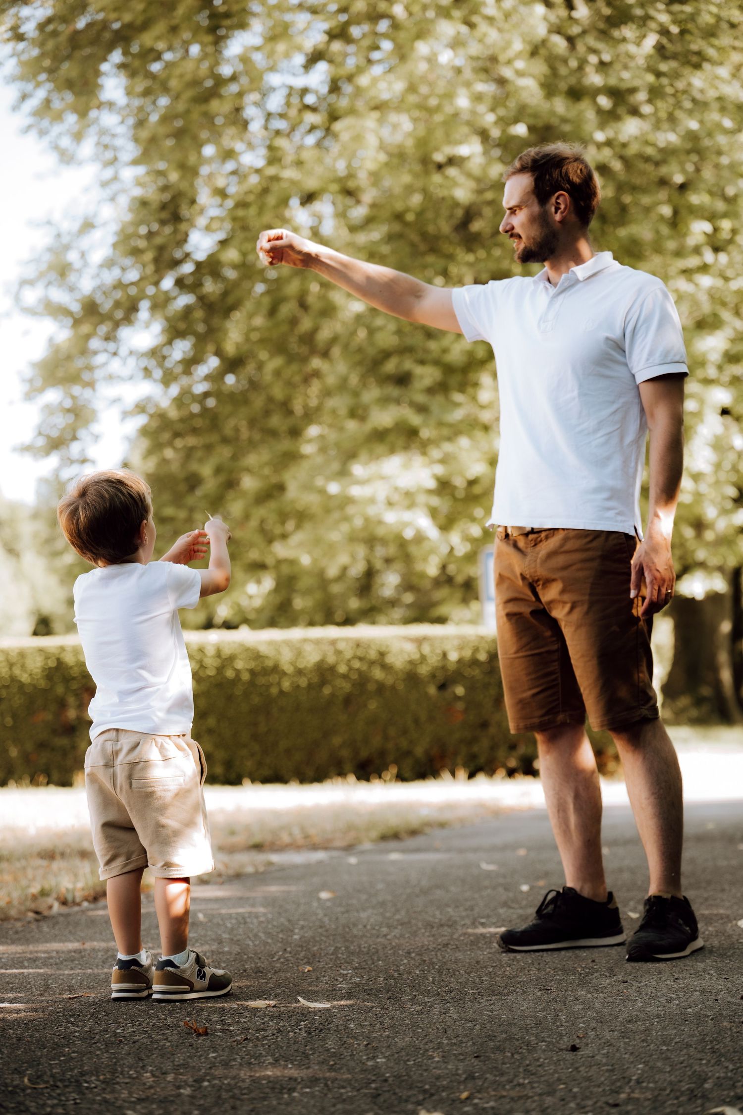 Father and young son playing together in a sunny park, both wearing white shirts and shorts on a tree-lined path.