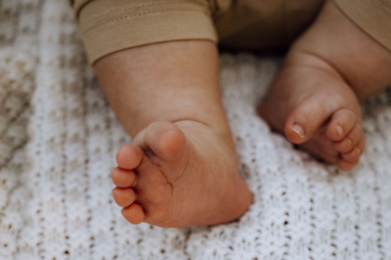 Close-up of tiny newborn baby feet resting on a soft white knit blanket, wearing beige onesie.