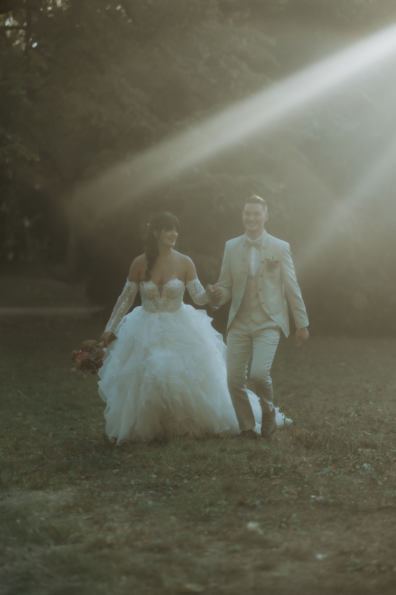 Bride and groom walk hand-in-hand through sunlit park, she in a ruffled white gown, he in a beige suit.