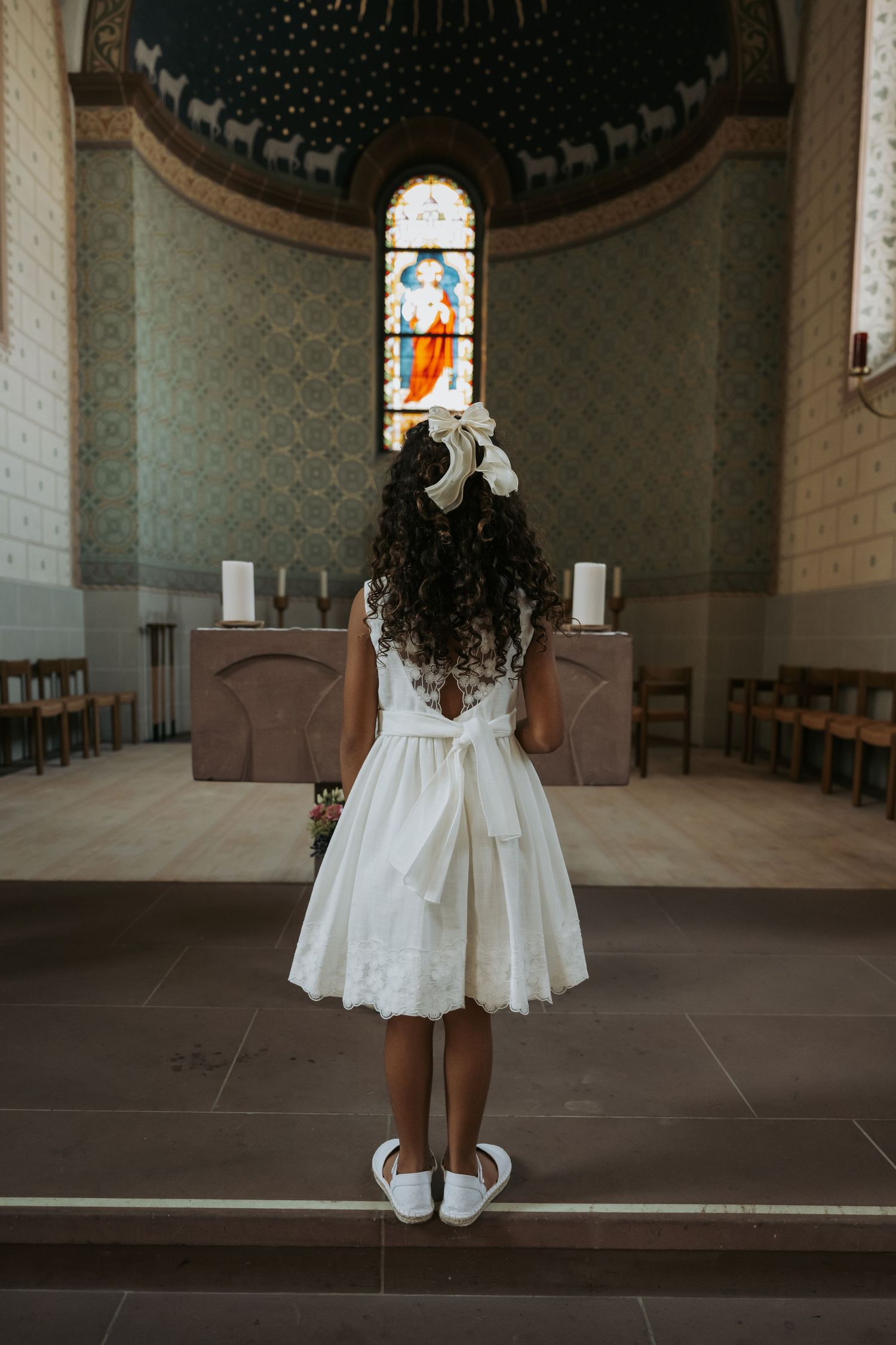 Young girl in white dress and bow stands before church altar with stained glass window during First Communion ceremony.