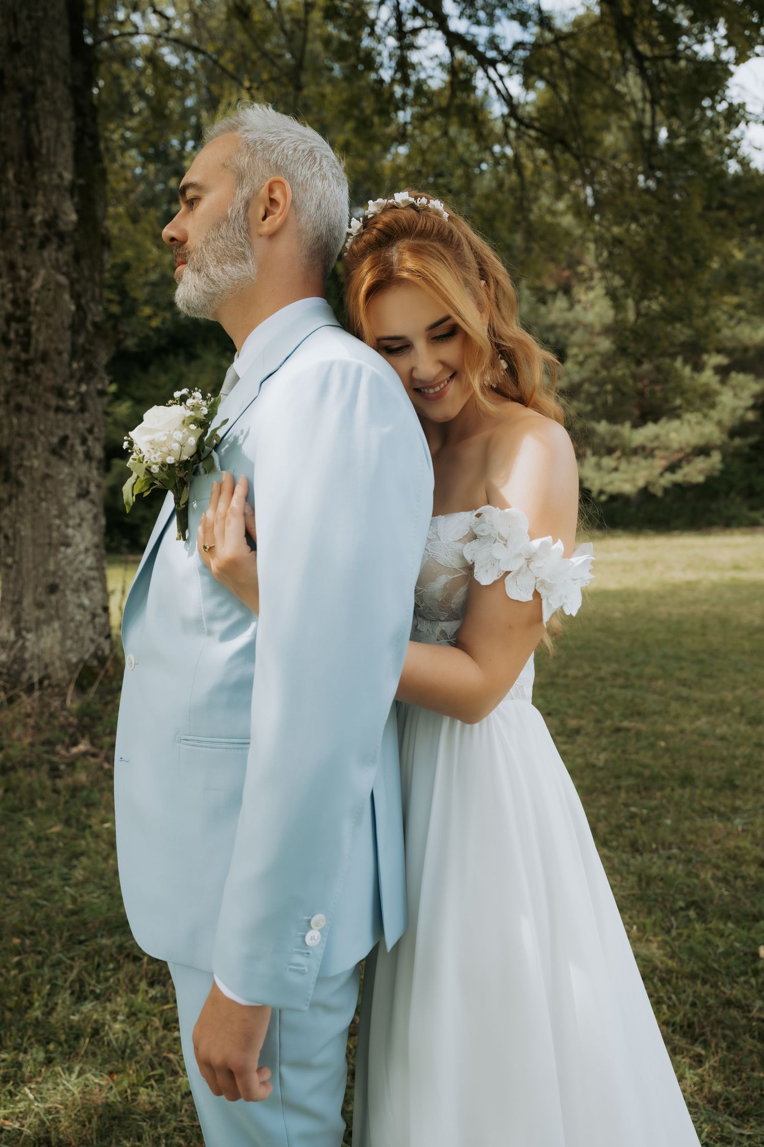 A smiling bride in a white floral gown embraces a groom in a light blue suit outdoors near a tree.