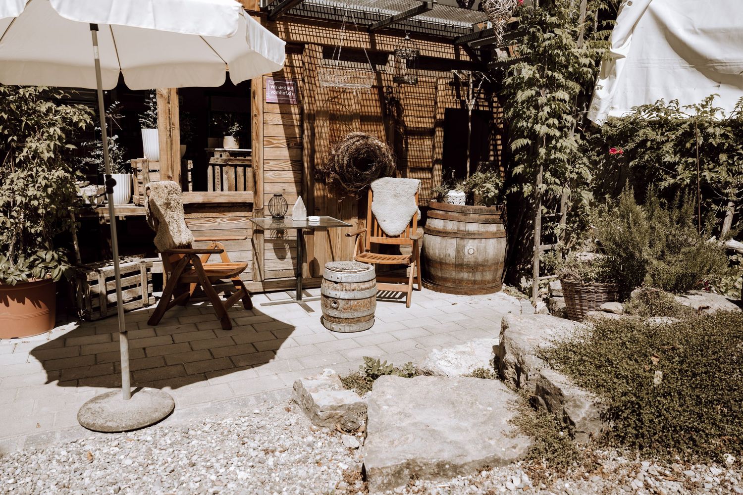 Rustic outdoor patio with wooden barrels, garden chairs, umbrella, and lush greenery surrounding a cozy wooden cabin.