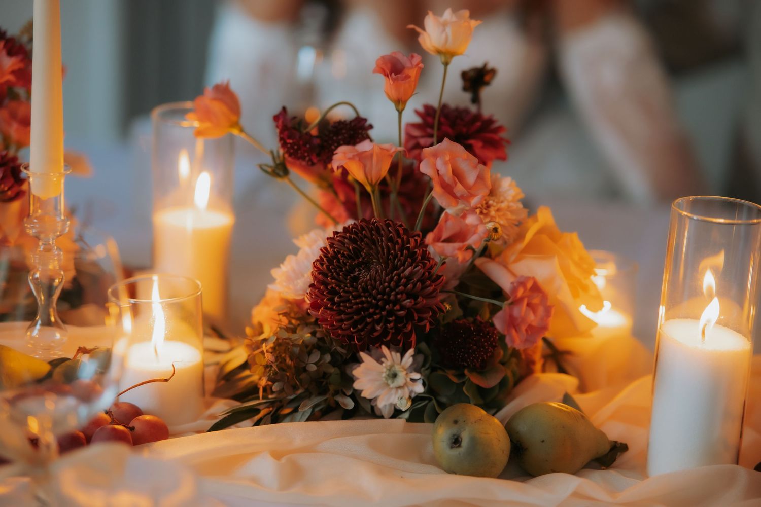 Autumn wedding table centerpiece with burgundy dahlias, coral flowers, candles, and pears on a draped white tablecloth.