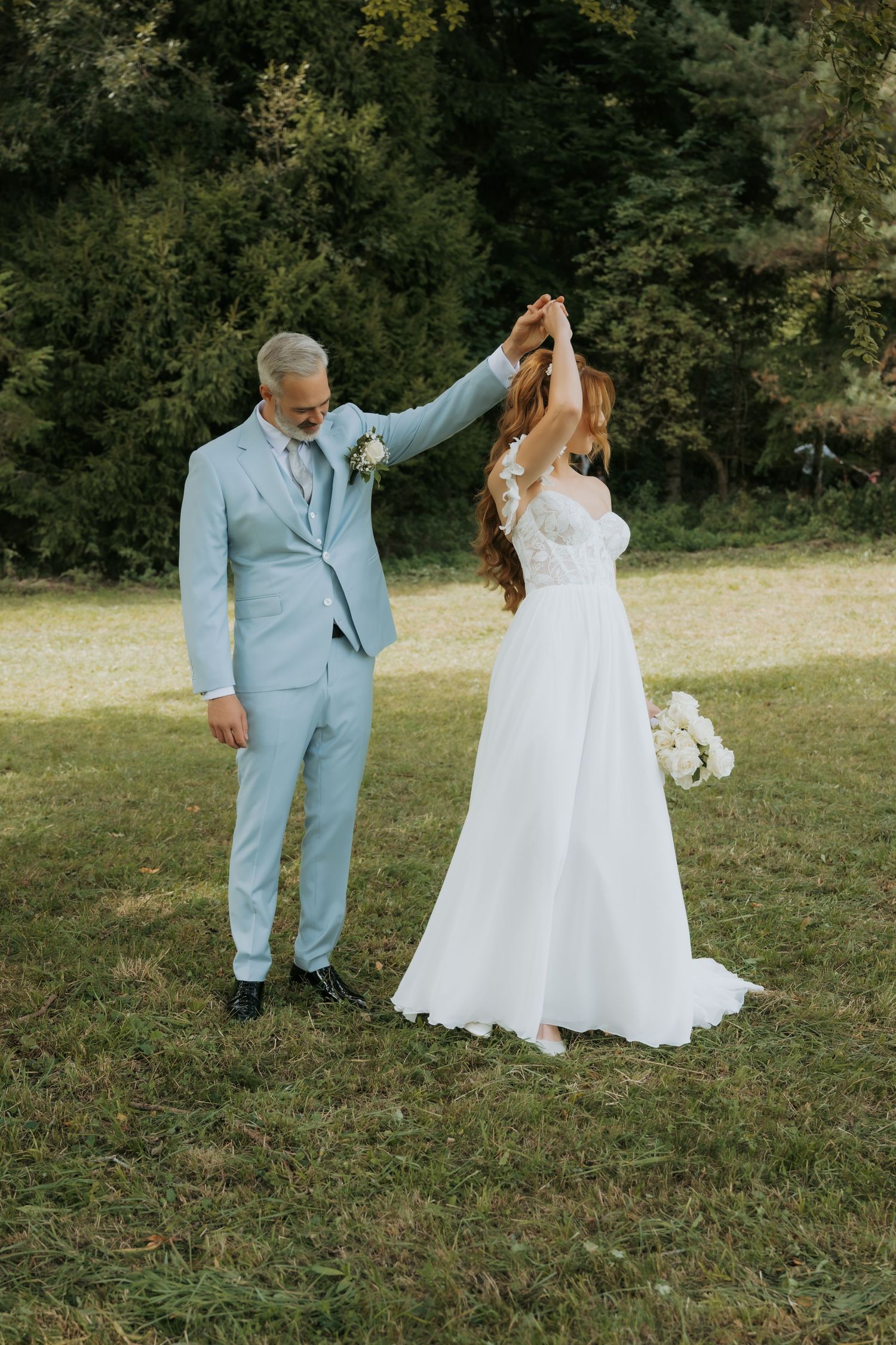 A groom in a light blue suit twirls his bride in a white wedding gown outdoors on a green lawn.