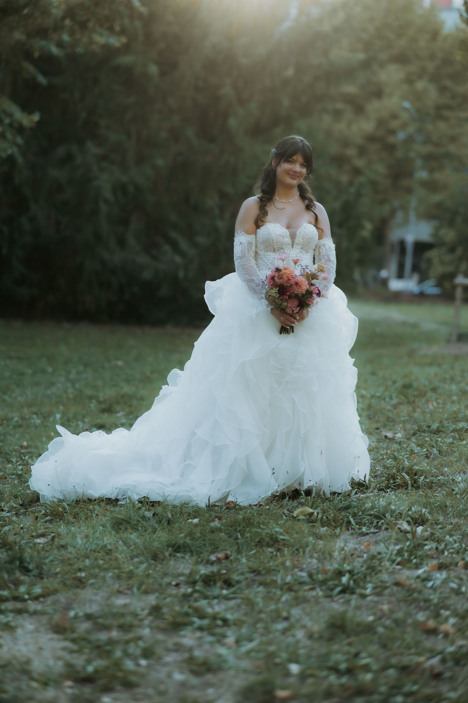 Bride in white off-shoulder ball gown holding colorful bouquet, standing on green grass with sunlit trees behind her.