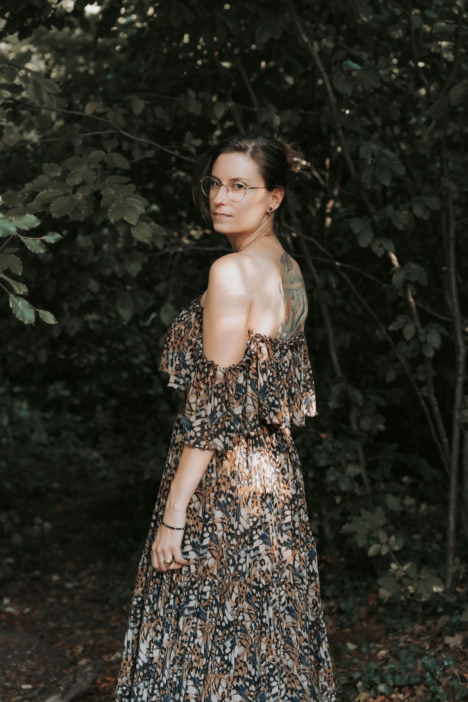 Woman in floral off-shoulder maxi dress and glasses poses gracefully among lush green trees in a forest setting.