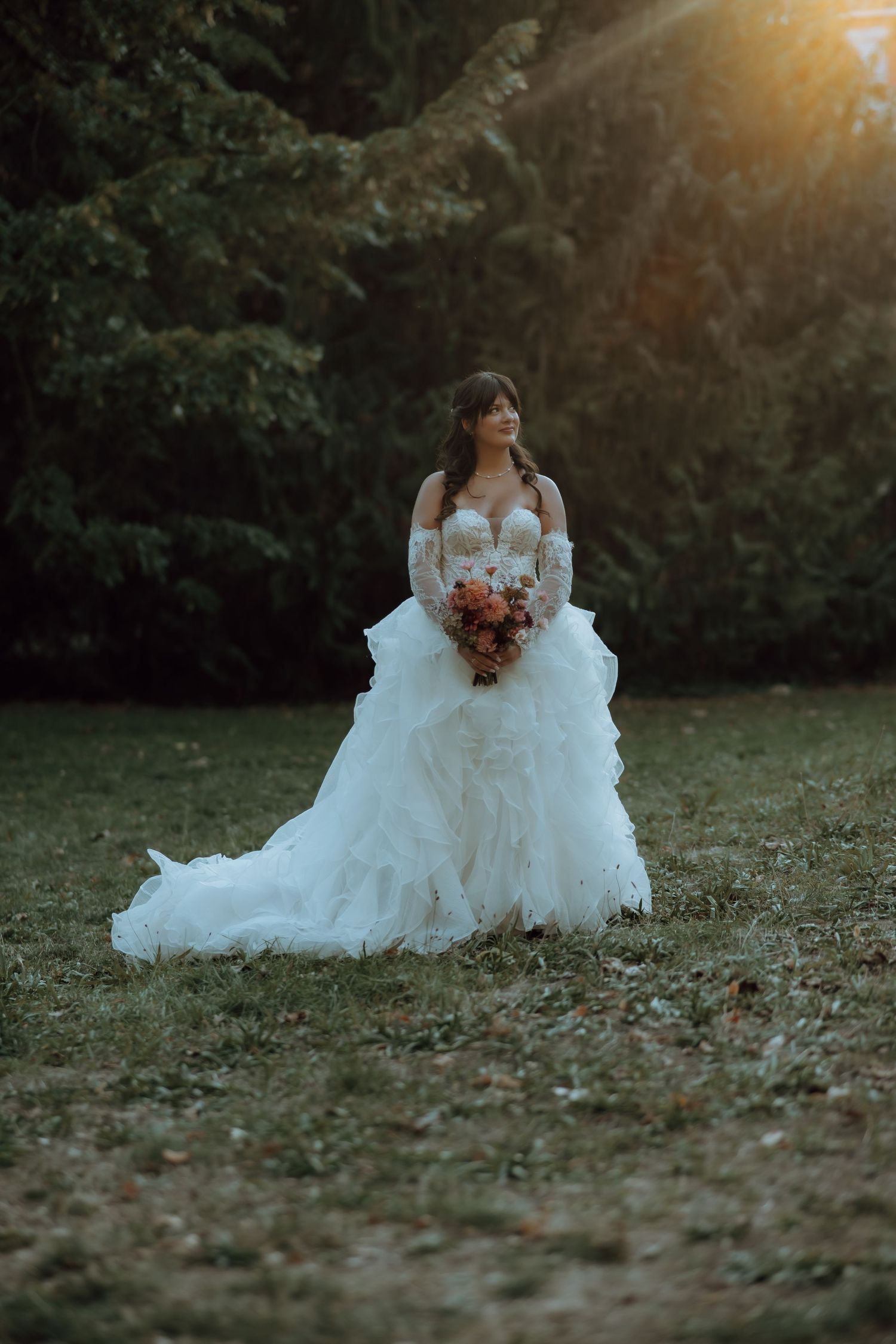 Bride in a white ruffled ball gown holding a bouquet, standing outdoors at golden hour with trees behind her.