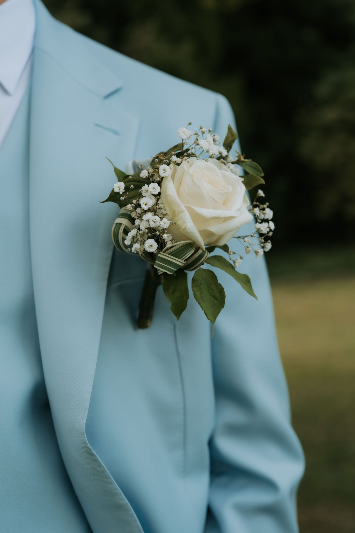 A white rose boutonniere with baby's breath and green ribbon on a light blue suit lapel.