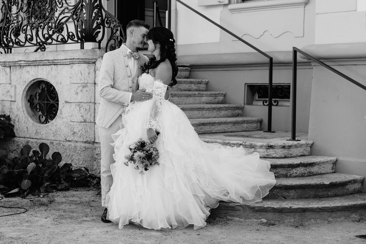 Black and white photo of a bride and groom sharing a romantic kiss on outdoor steps, bride holding a bouquet.
