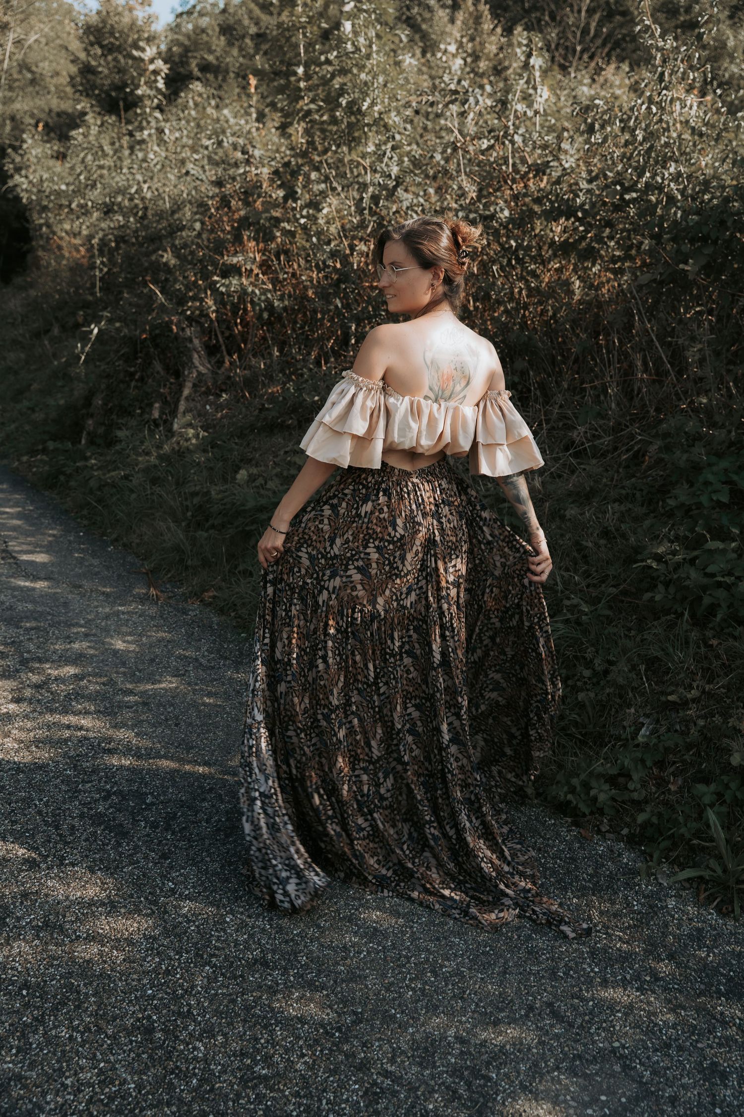 Woman in off-shoulder crop top and floral maxi skirt standing on a path surrounded by lush greenery.