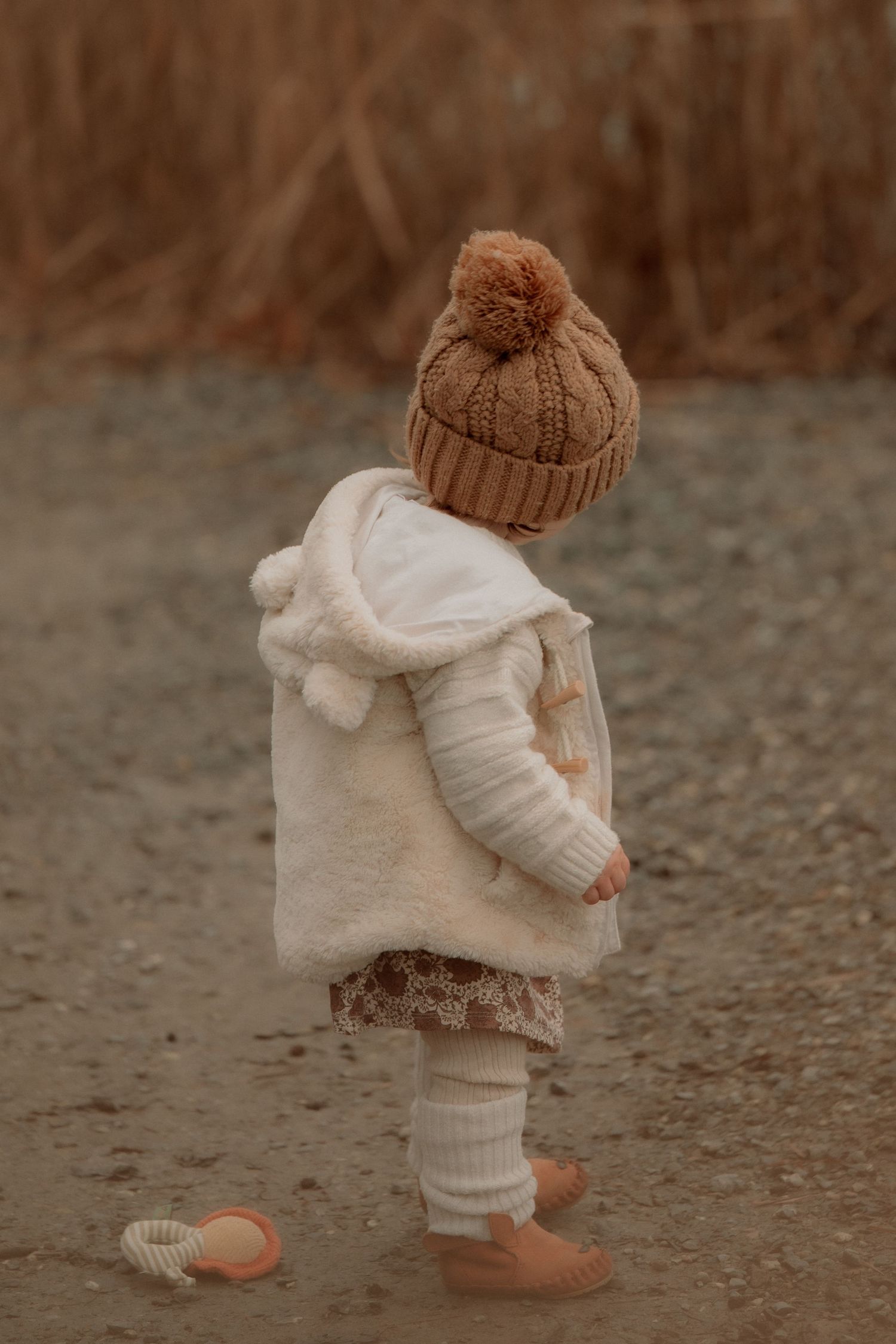 Toddler in cream coat, knit pom-pom hat, and white tights standing outdoors in autumn setting.