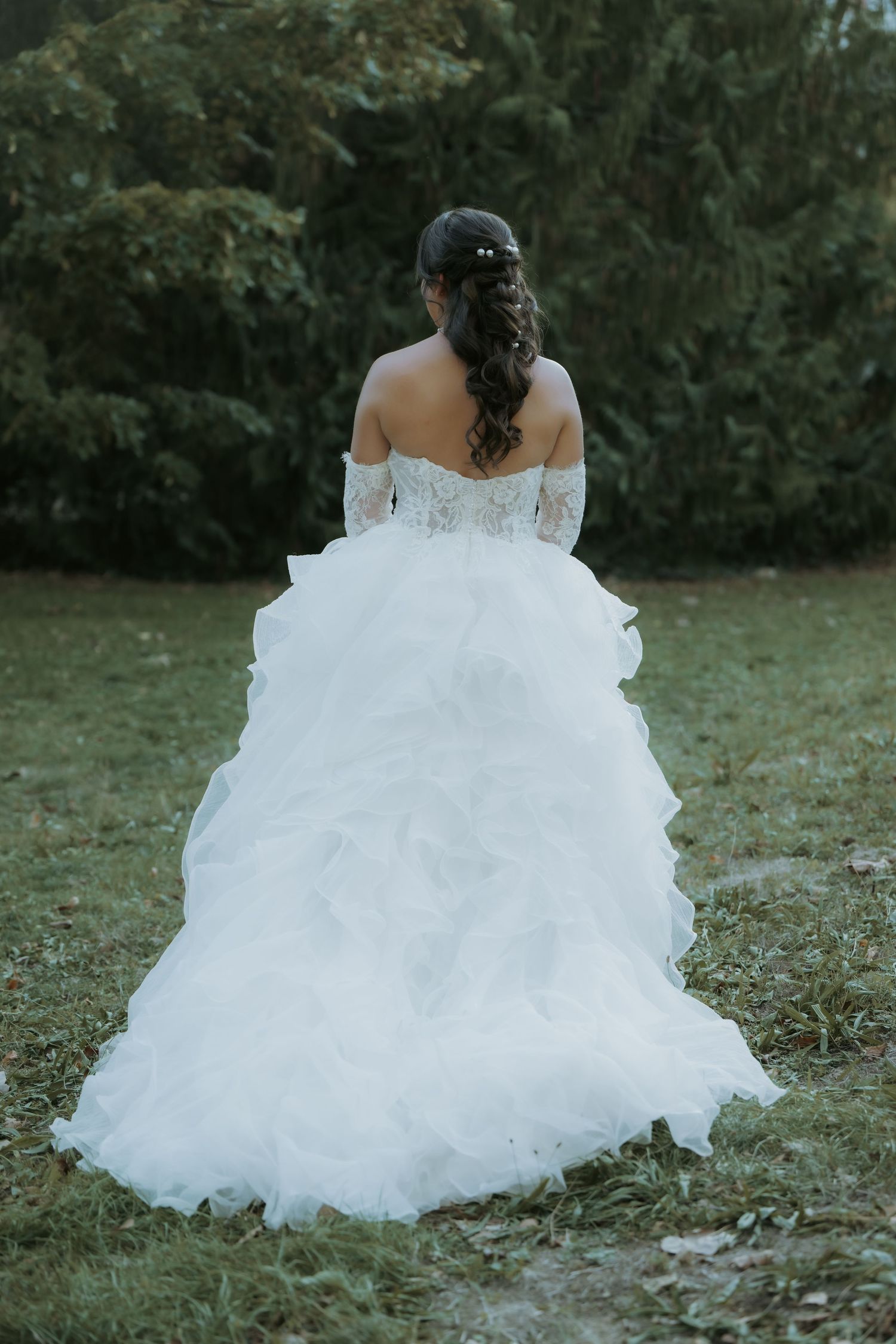Bride in white off-shoulder ballgown with lace bodice and ruffled skirt, standing with back to camera in garden.