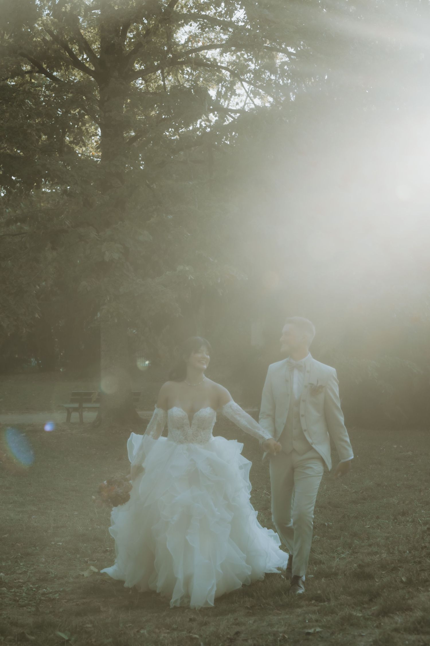 Bride and groom walking hand in hand through a sunlit park, bride in ruffled white gown, groom in beige suit.
