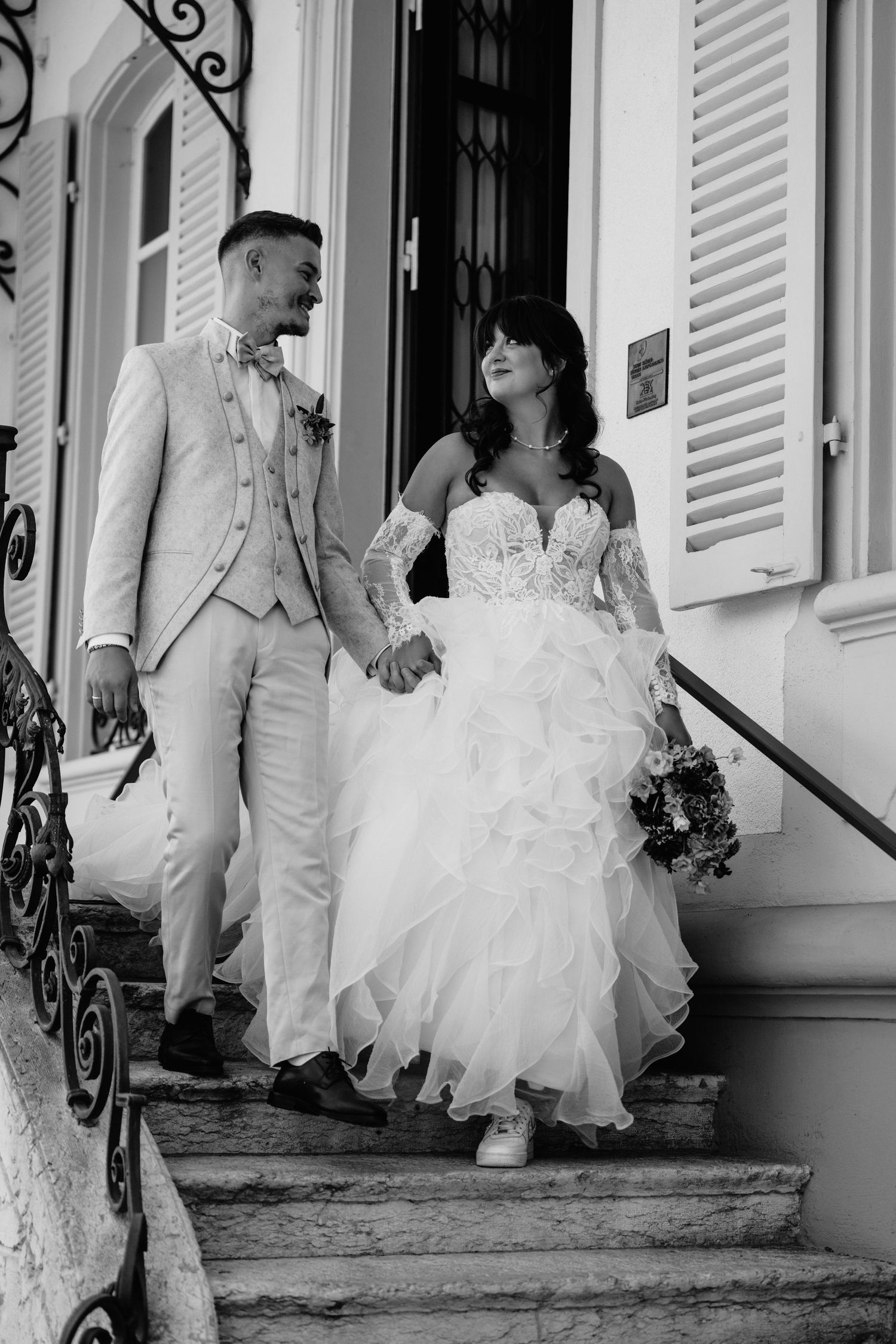 Black and white photo of a bride and groom holding hands, walking down stone steps outside an elegant building.