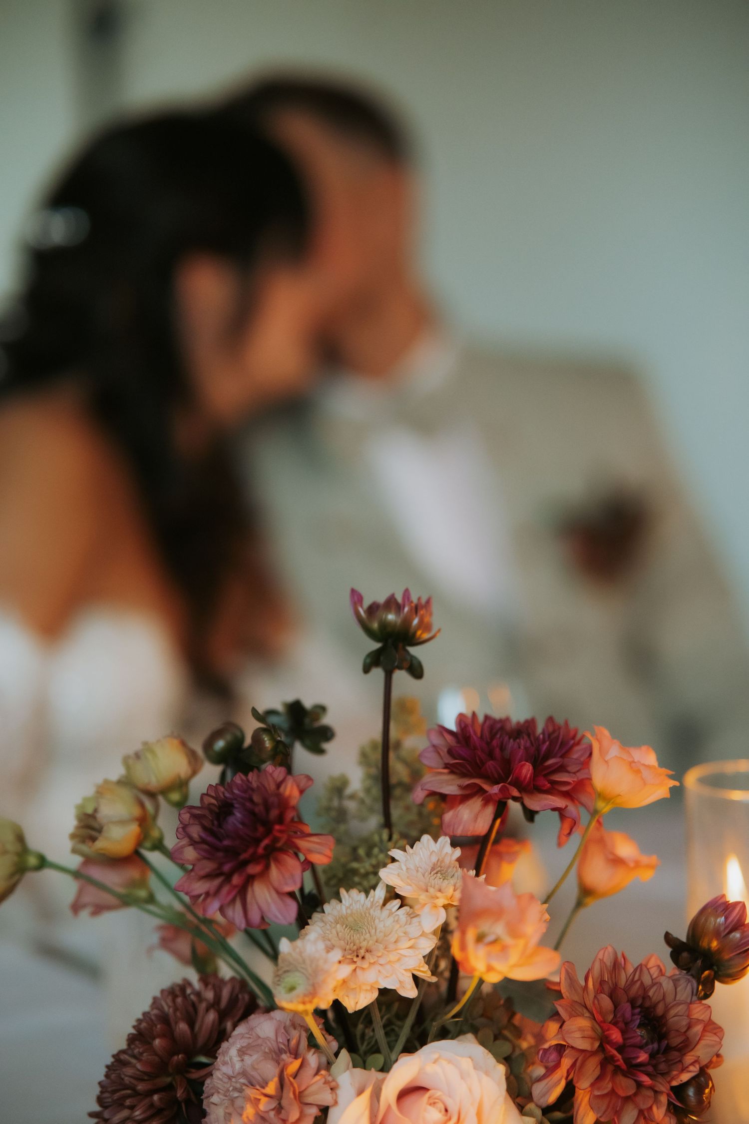 Blurred bride and groom kissing behind a vibrant floral centerpiece of dahlias and roses with a candle at a wedding reception.