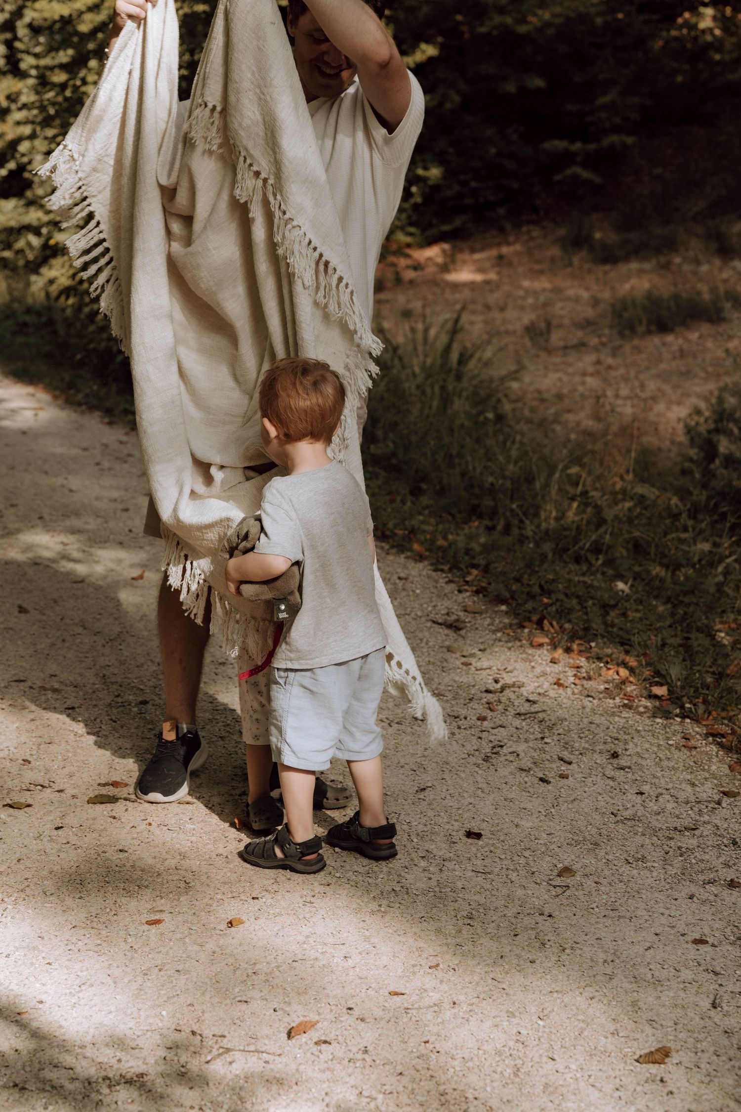 A toddler with red hair stands on a dirt path next to an adult holding a cream fringed blanket outdoors.