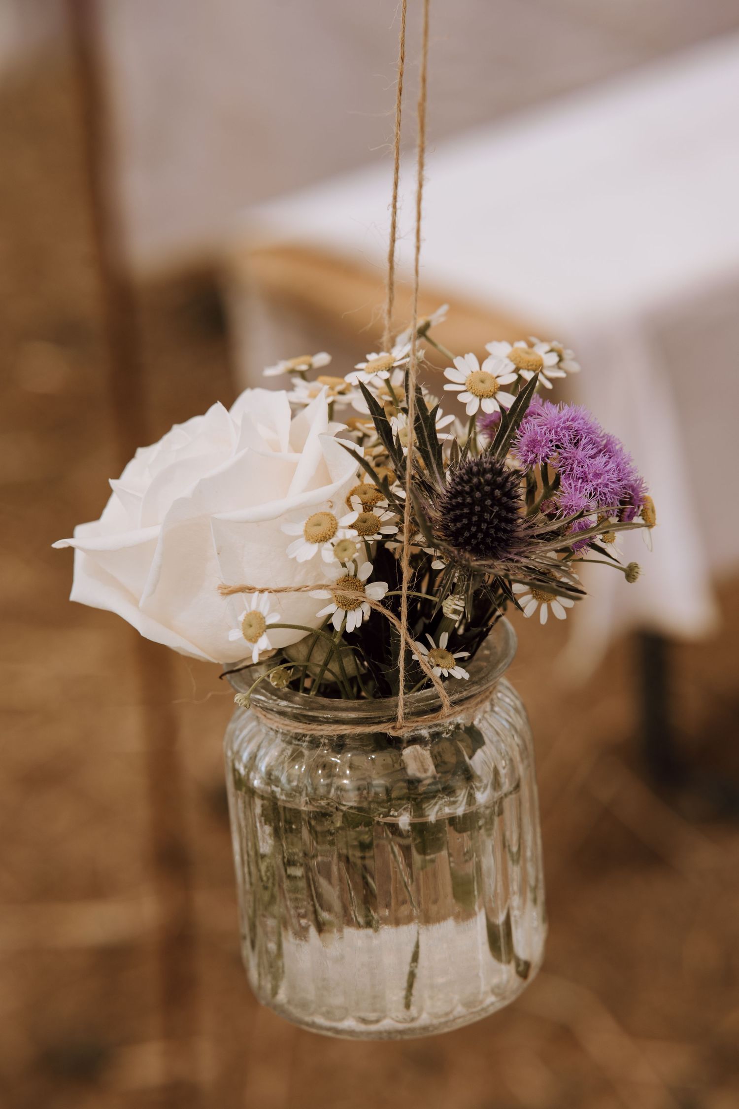 A rustic mason jar with white roses, daisies, and purple thistle flowers hanging by twine string.