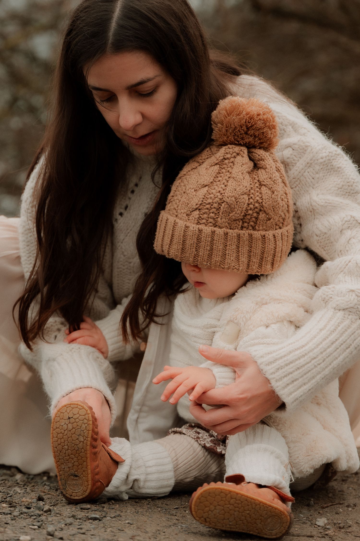 A mother in white knitwear helps her baby in a brown pom-pom hat and moccasins sit outdoors.