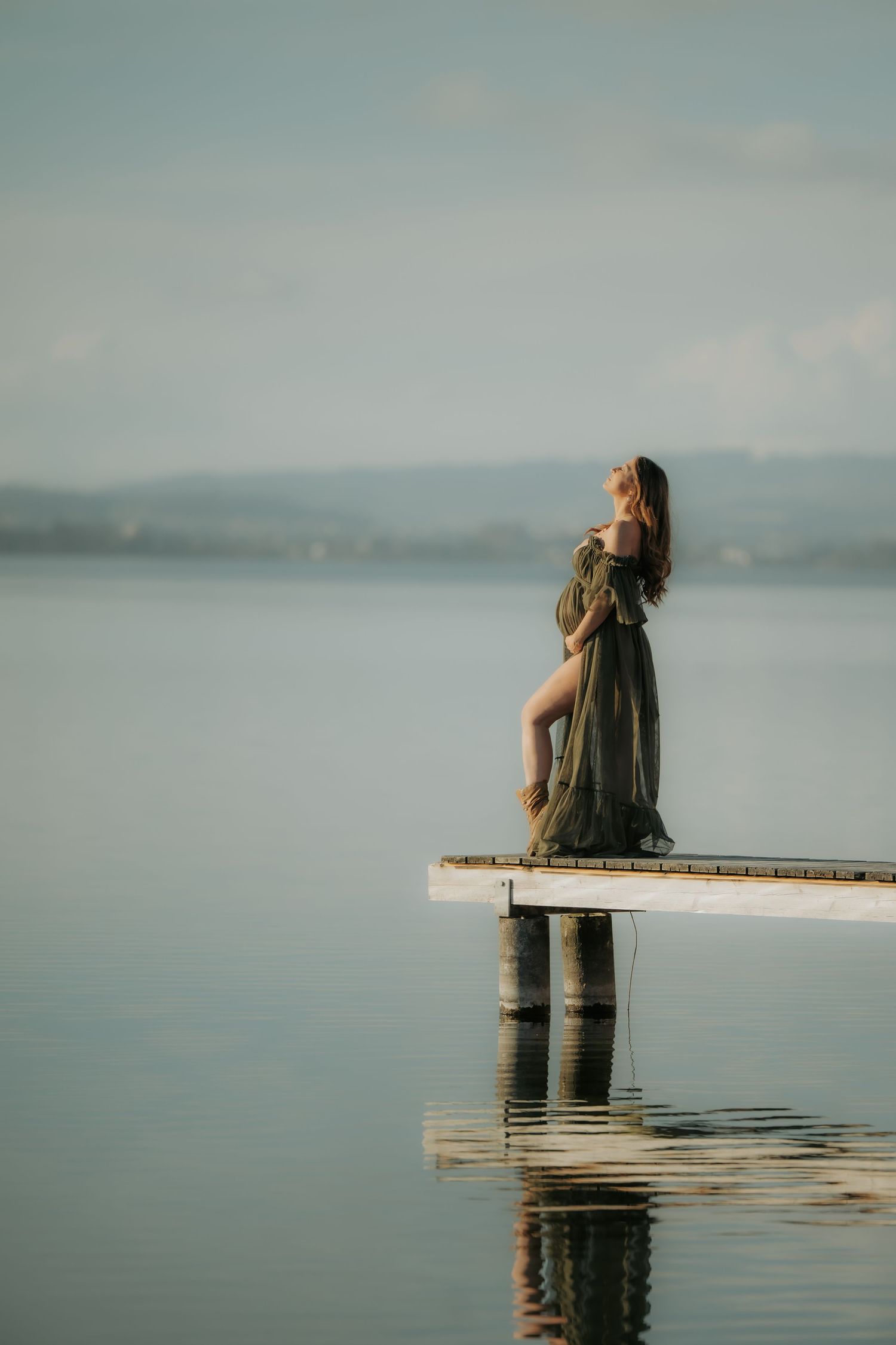 Pregnant woman in flowing olive dress standing on a wooden dock over still lake.